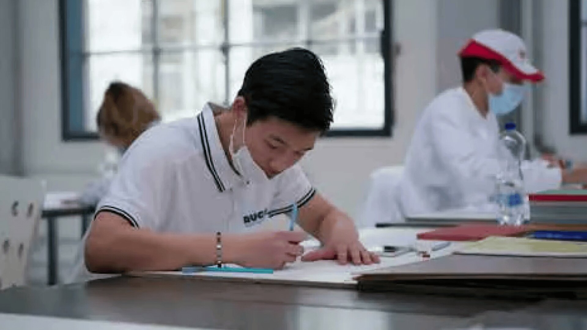 Ein Student mit Gesichtsmaske und weißem Poloshirt sitzt an einem Schreibtisch und schreibt in ein Notizbuch. Andere Personen, ebenfalls maskiert, sitzen und arbeiten im Hintergrund.
