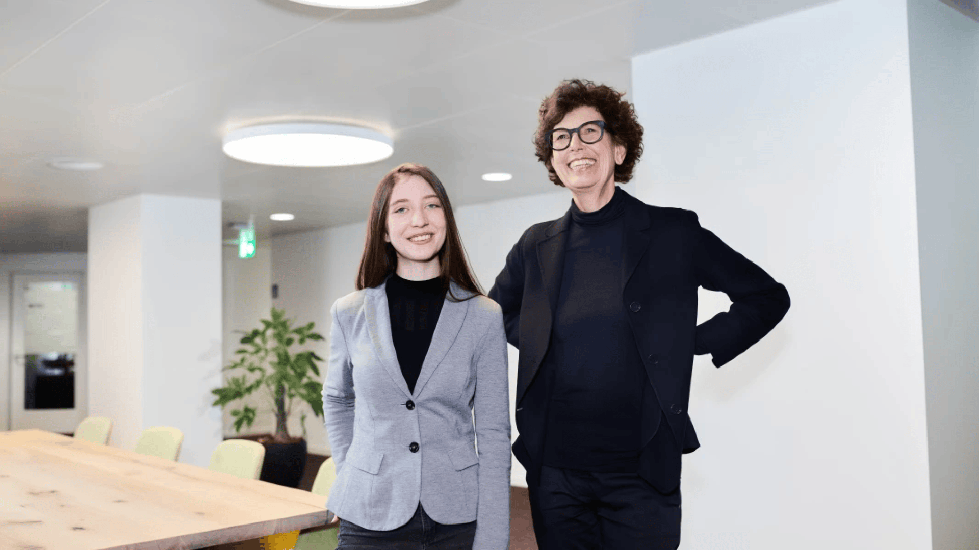Zwei Frauen in Business-Kleidung stehen lächelnd in einem modernen Büroraum mit einem langen Holztisch, Stühlen und einer Pflanze im Hintergrund. Die Umgebung ist hell mit weißen Wänden und Deckenleuchten.