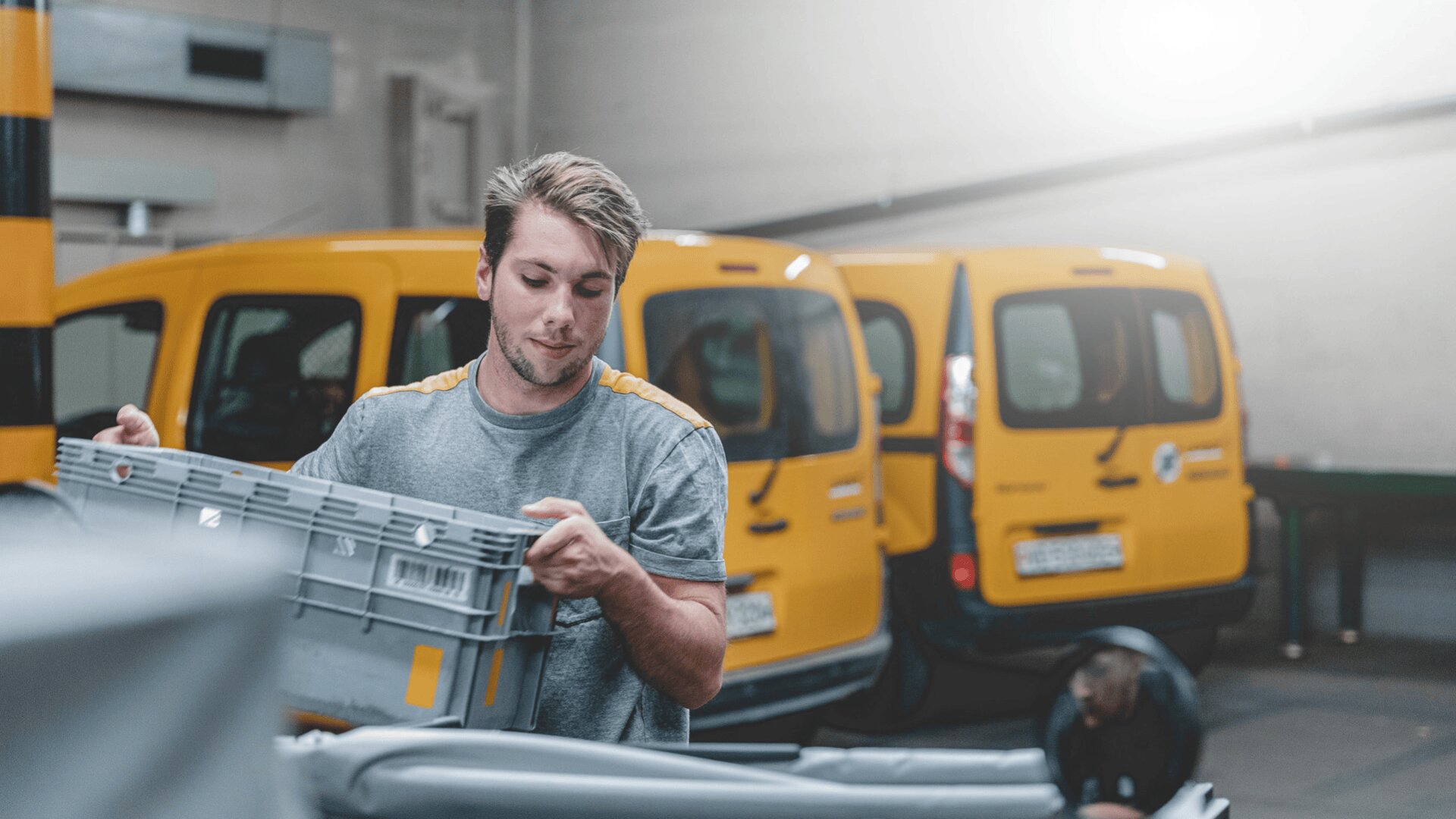 Ein Zusteller sortiert Pakete in einer Lagerhalle und hält eine graue Plastikkiste in der Hand. Im Hintergrund sind zwei gelbe Lieferwagen geparkt, die hell beleuchtet sind.