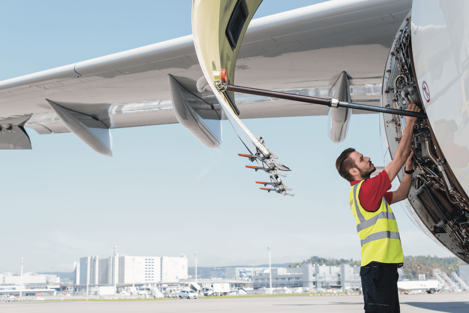 Ein männlicher Flughafenarbeiter in einer gelben Sicherheitsweste inspiziert oder repariert das Triebwerk eines Flugzeugs mit geöffneter Triebwerksabdeckung auf dem Rollfeld eines Flughafens, wobei im Hintergrund moderne Gebäude zu sehen sind.