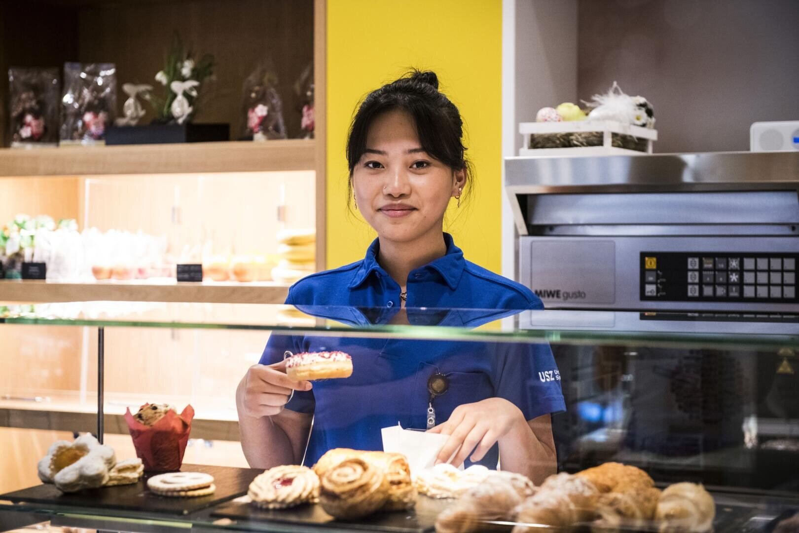 Eine Frau in einem blauen Hemd steht hinter einer Bäckertheke und hält einen Donut mit rosa Zuckerguss in der Hand. Vor ihr sind Gebäckstücke ausgestellt, und im Hintergrund stehen Regale mit weiteren Leckereien. Sie lächelt leicht.