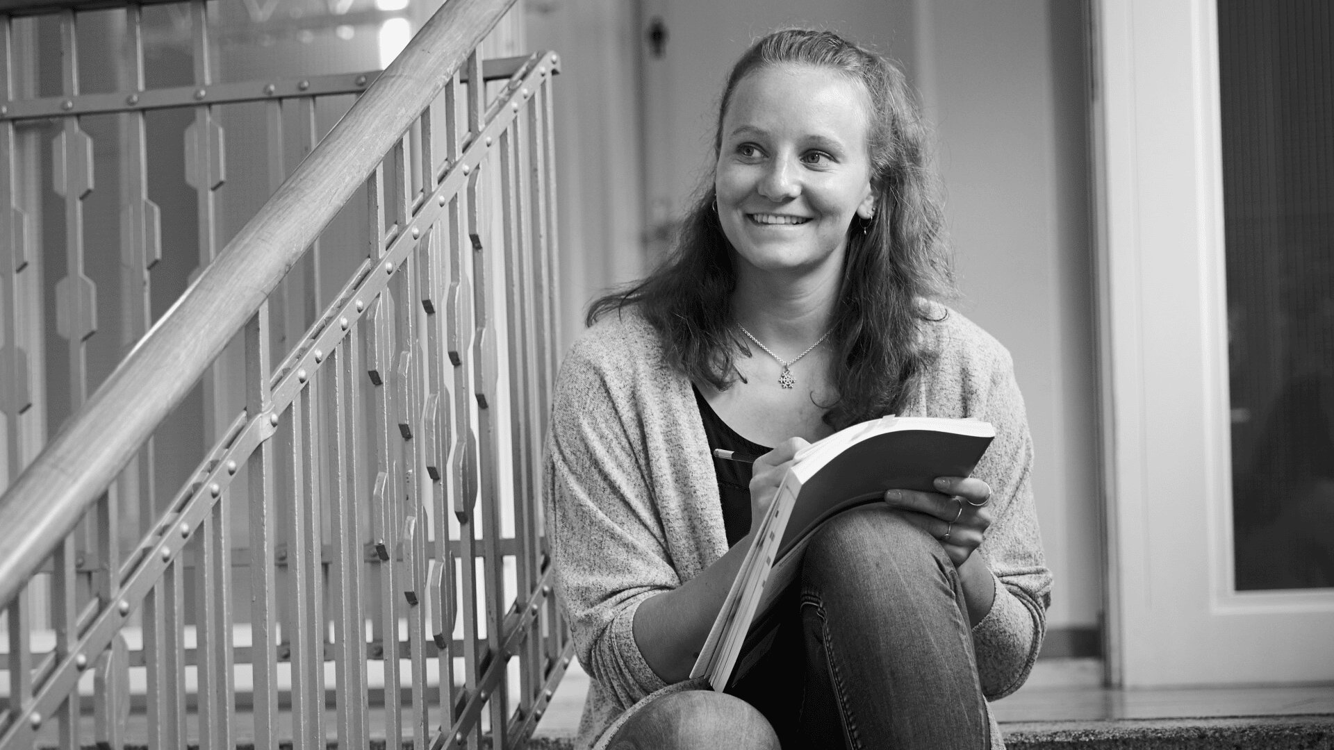 Eine junge Frau sitzt auf einer Innentreppe, lächelt und hält ein Notizbuch in der Hand, während sie schreibt. Sie hat lange Haare und ist leger gekleidet, neben ihr steht ein Treppengeländer. Das Bild ist in schwarz-weiß gehalten.