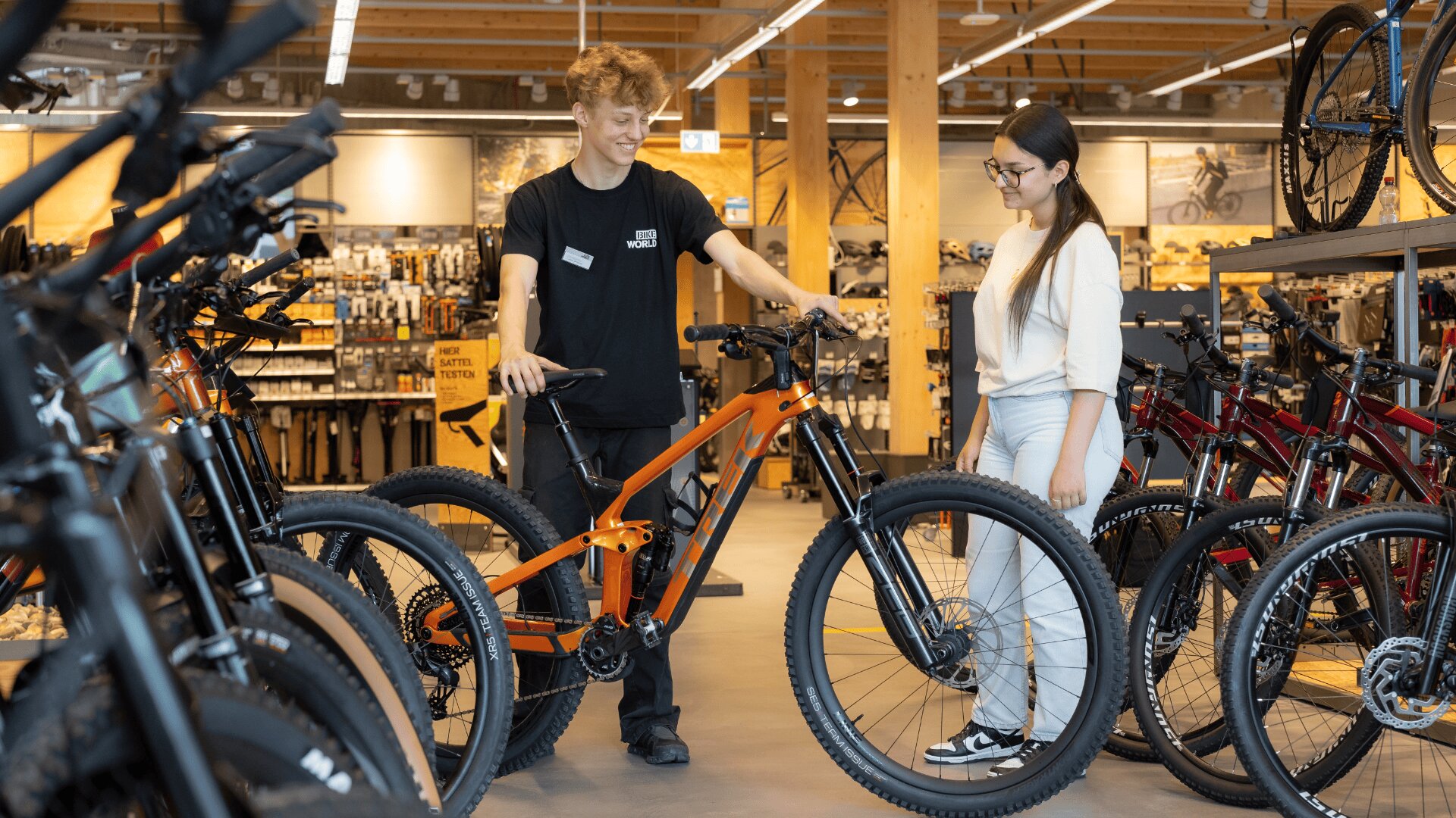 Ein Angestellter zeigt einem lächelnden Kunden in einem Fahrradgeschäft ein orangefarbenes Mountainbike, umgeben von verschiedenen anderen ausgestellten Fahrrädern und Zubehör.