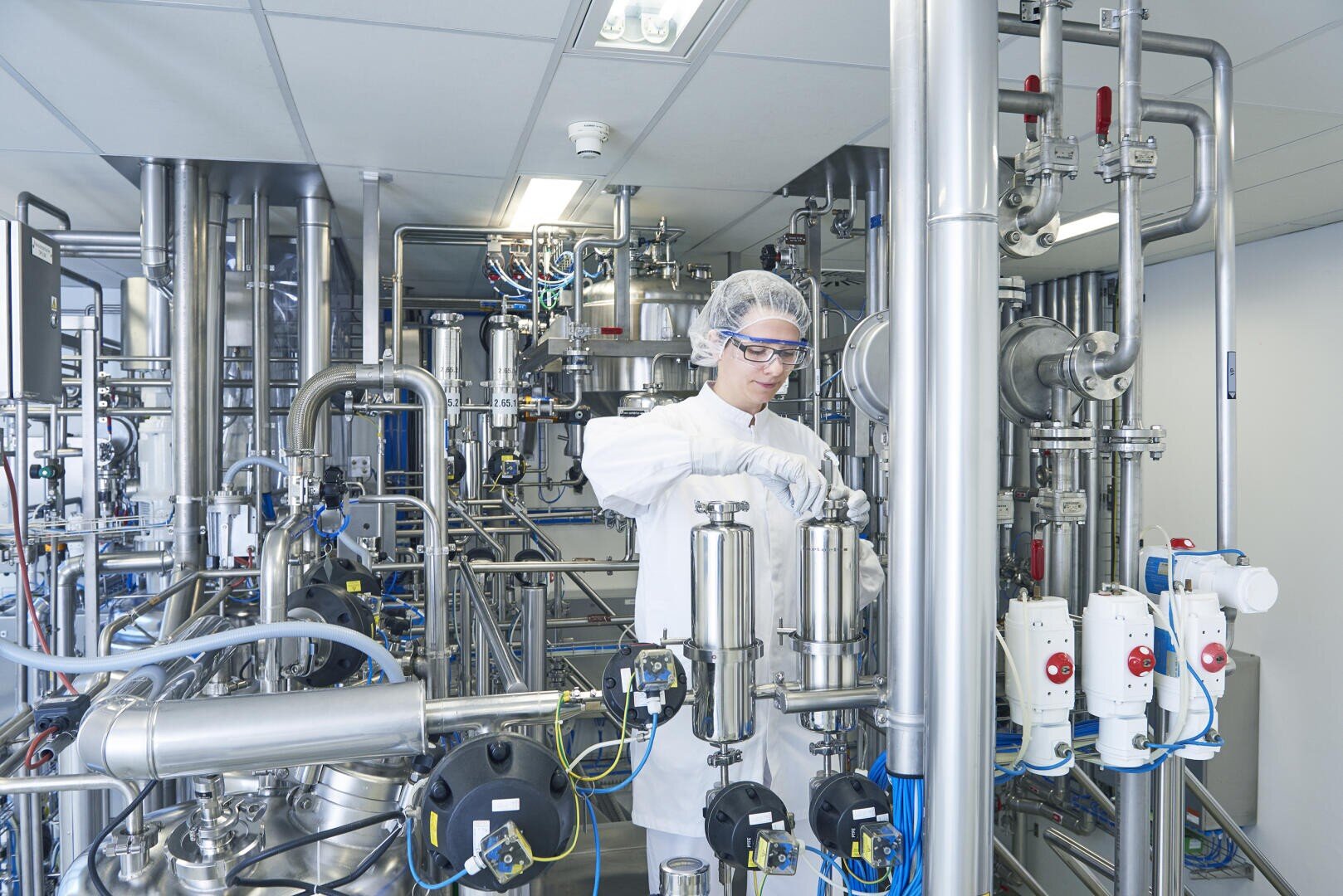 Eine Person in Schutzkleidung, Haarnetz und Schutzbrille bedient Maschinen in einem sauberen Industrielabor, das mit Metallrohren, Tanks und Steuergeräten gefüllt ist.