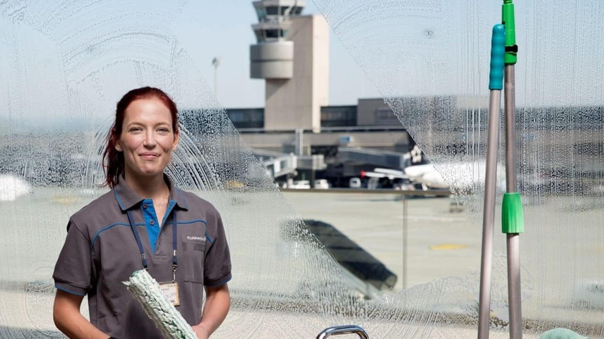 Eine Frau in einer Reinigungsuniform steht vor einem großen, seifigen Fenster auf einem Flughafen. Sie hält ein Fensterputzgerät in der Hand, während im Hintergrund der Kontrollturm des Flughafens und Flugzeuge zu sehen sind.
