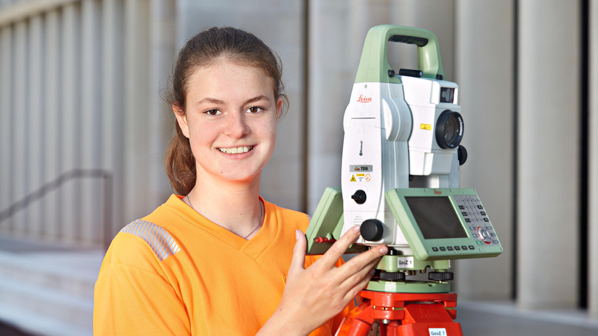 Eine junge Frau in einem orangefarbenen Hemd lächelt, während sie ein grün-weißes Vermessungsinstrument auf einem Stativ bedient, während im Hintergrund ein unscharfes Gebäude zu sehen ist.
