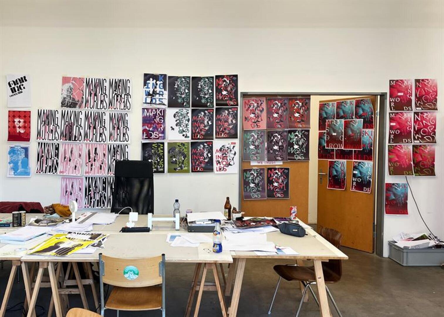 Künstleratelier mit bunten Plakaten an den Wänden, Papieren und Kunstzubehör auf Holztischen und -stühlen und einem schwarzen Bürostuhl vor den Plakaten. Der Raum wirkt geschäftig und kreativ.