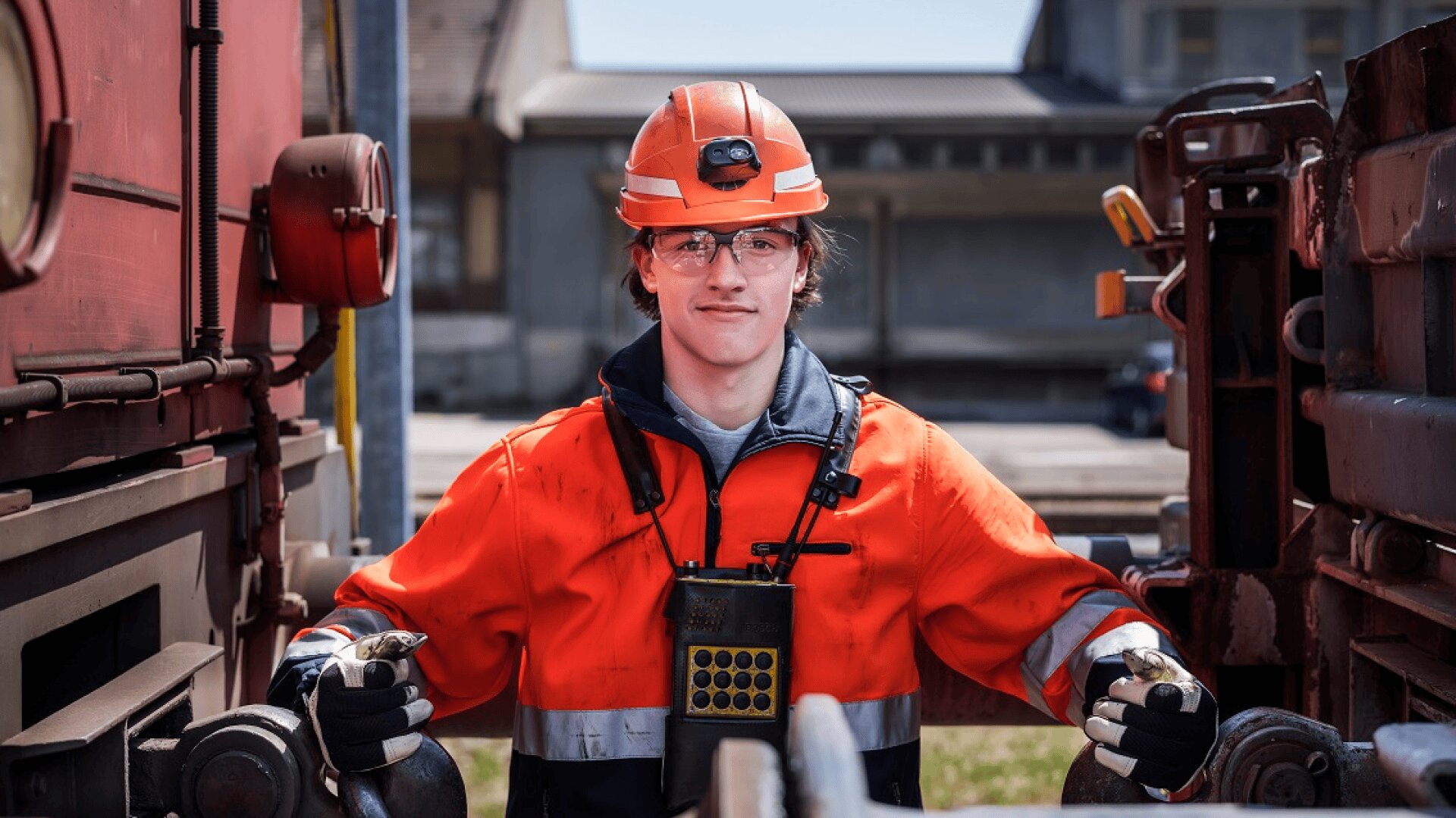 Ein Eisenbahner in einer orangefarbenen Sicherheitsuniform, mit Helm und Brille steht an einem sonnigen Tag zwischen zwei Zugwaggons und betätigt im Freien eine Kupplungsausrüstung.