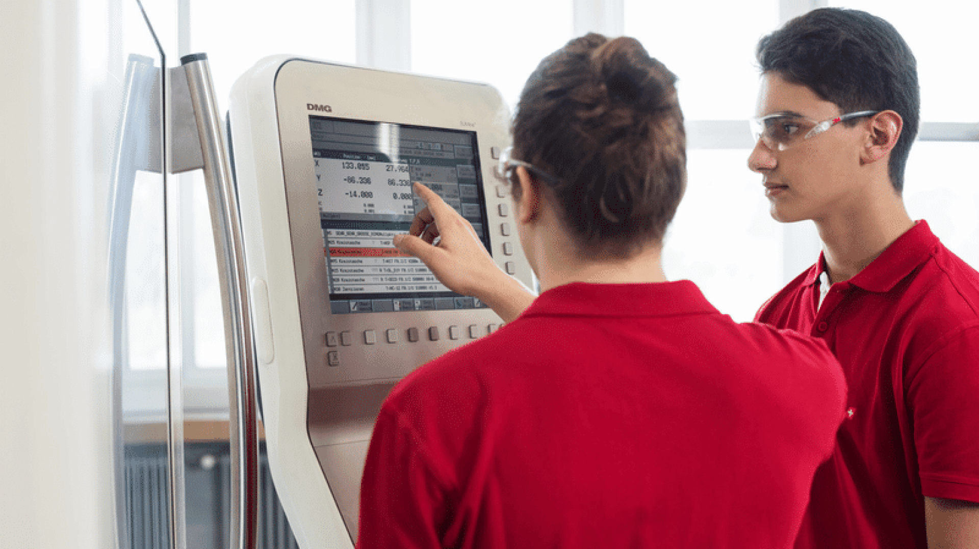 Zwei Personen in roten Hemden, von denen eine auf ein Touchscreen-Display mit Daten und Diagrammen zeigt, während die andere sie beobachtet. Sie stehen in einem Innenraum, beide auf den Bildschirm konzentriert, und einer trägt eine Schutzbrille.