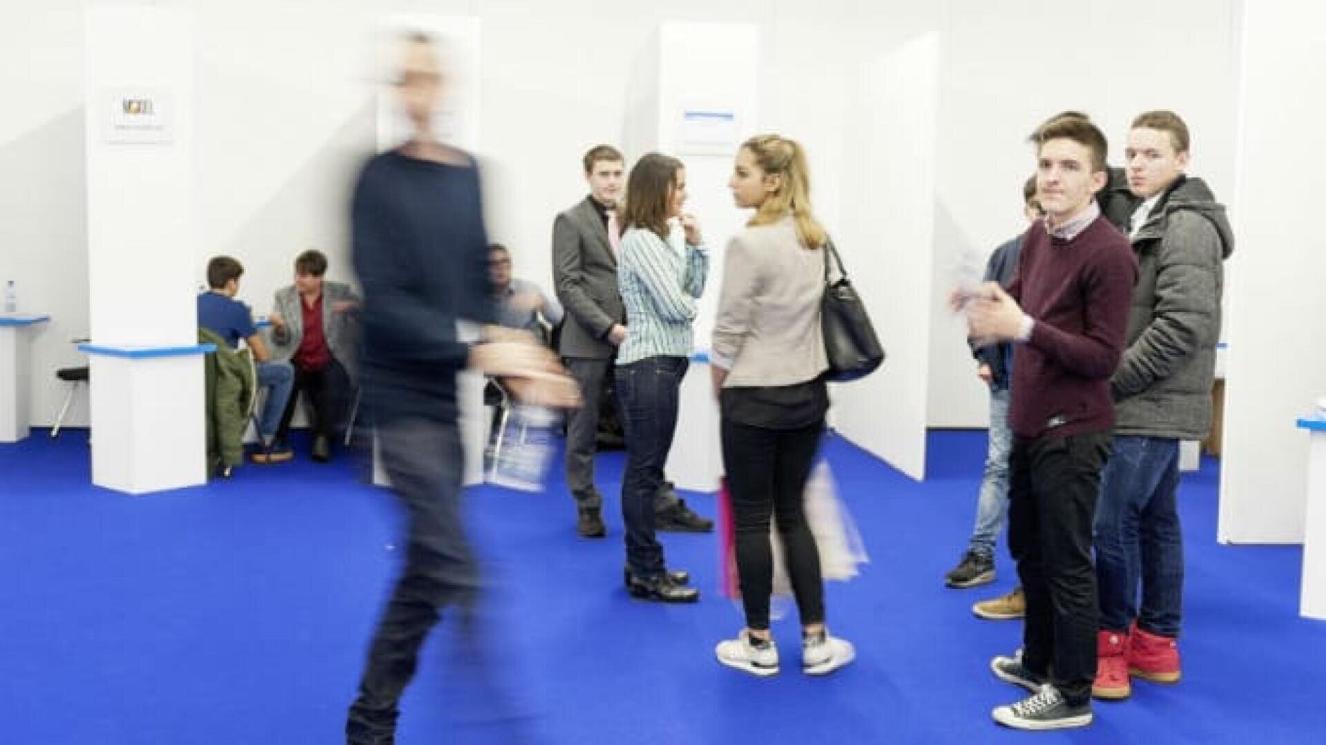 Menschen stehen und sitzen in einem mit blauem Teppich ausgelegten Raum mit weißen Kabinen. Eine Person geht vorbei, verschwommen in der Bewegung, während mehrere andere warten oder sich unterhalten, einige schauen in die Kamera.