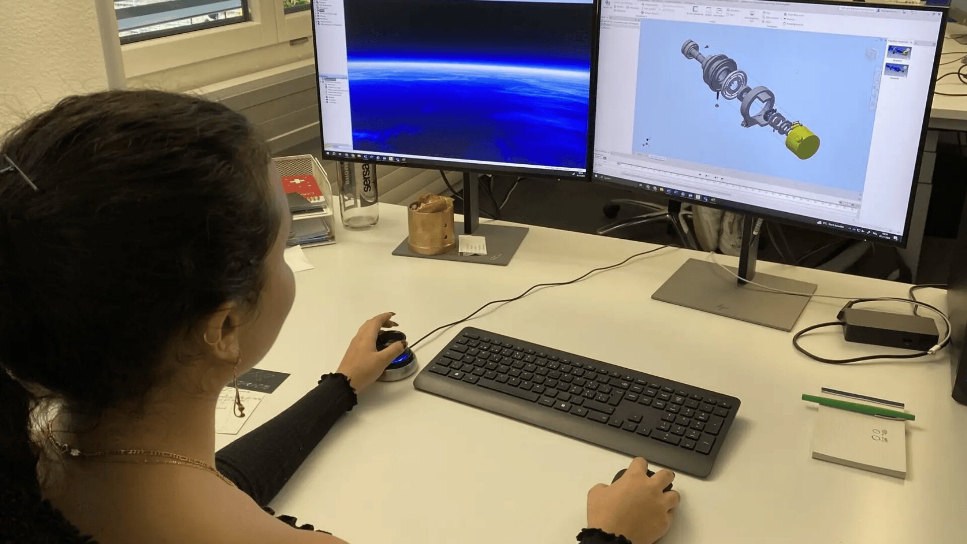 Eine Person sitzt an einem Schreibtisch mit einem Computer mit zwei Monitoren; auf einem Bildschirm ist ein mechanisches 3D-Modell zu sehen, auf dem anderen ein Bild der Erde aus dem Weltraum. Auf dem Schreibtisch befinden sich eine Tastatur, eine Maus und Büromaterial.