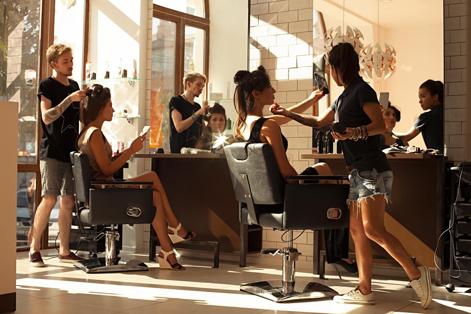 Zwei Frauen sitzen in den Stühlen des Salons und lassen sich von zwei Stylisten die Haare und das Make-up machen. Große Fenster lassen natürliches Licht herein und spiegeln die geschäftige, moderne Atmosphäre des Salons wider. Im Hintergrund befinden sich Regale mit Produkten und Spiegeln.