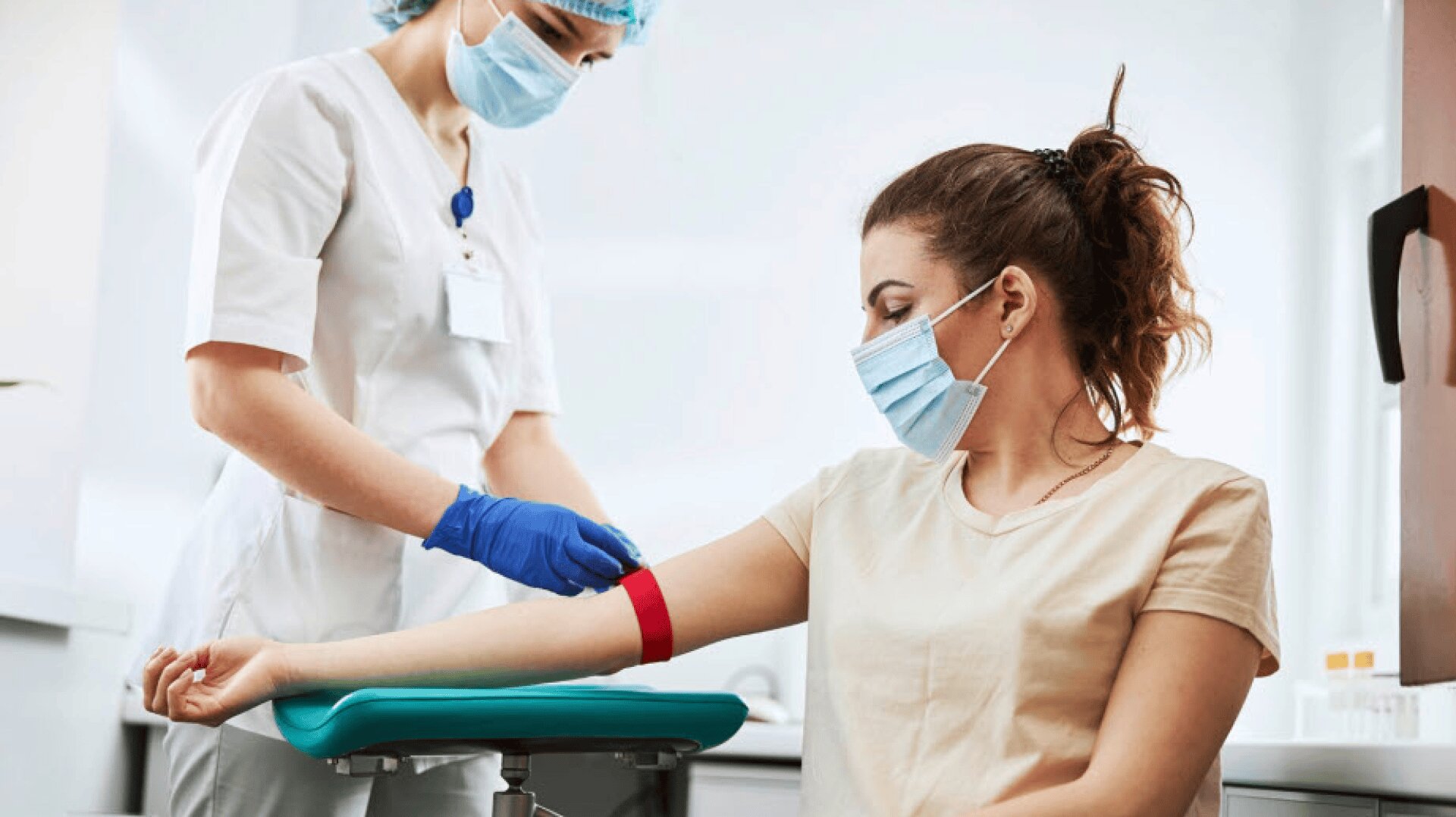 Eine medizinische Fachkraft mit Handschuhen und Maske bereitet sich darauf vor, einer sitzenden Frau mit Maske Blut abzunehmen, indem sie eine rote Aderpresse an ihrem Arm anlegt.