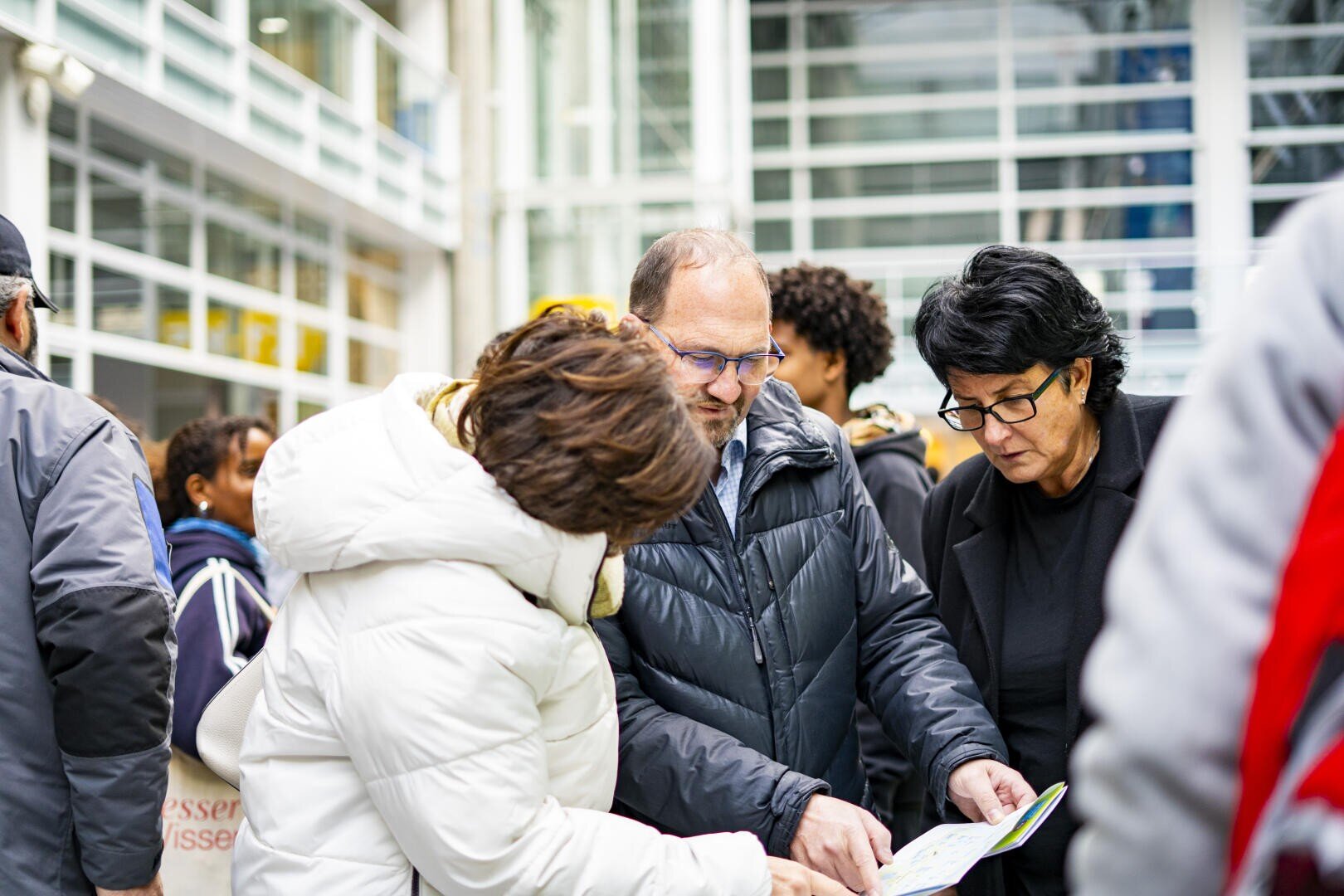 Drei Erwachsene in Wintermänteln stehen eng beieinander in einem belebten Innenraum und prüfen eine Karte oder ein Papier. Im Hintergrund sind andere Personen zu sehen, und Glaswände lassen natürliches Licht herein.