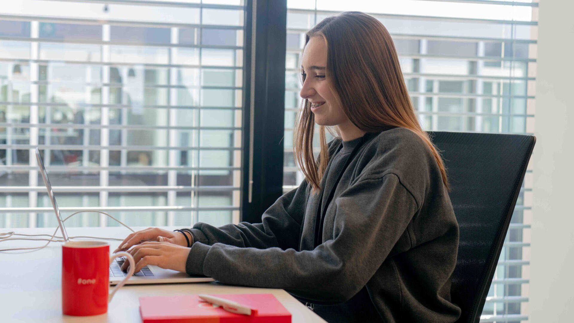 Eine junge Frau mit langen braunen Haaren sitzt an einem Schreibtisch und tippt auf einem Laptop. Sie lächelt und trägt ein dunkles Sweatshirt. Auf dem Schreibtisch stehen eine rote Tasse, ein Notizbuch und ein Stift. Im Hintergrund sind große Fenster mit Jalousien zu sehen.