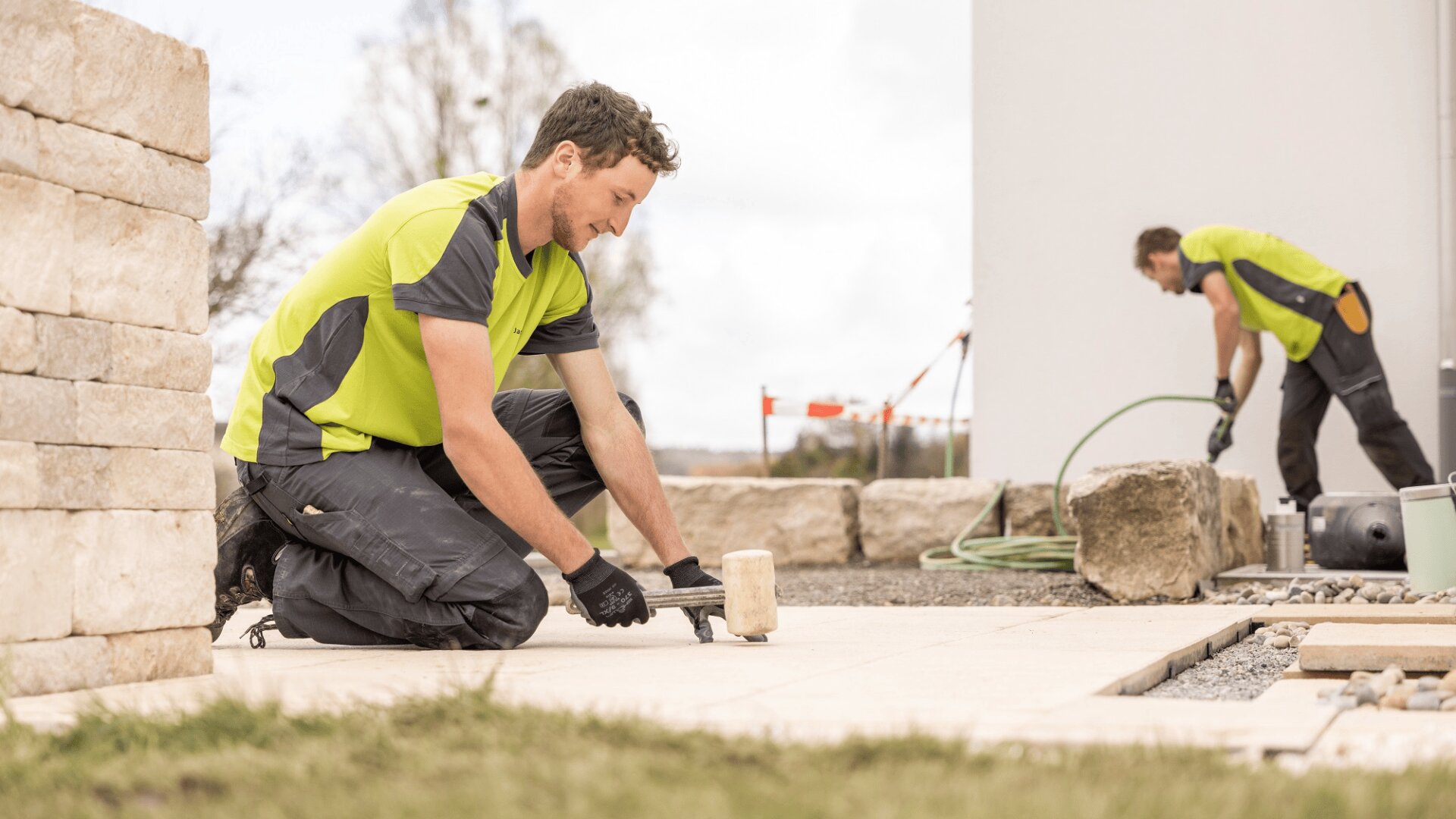 Zwei Bauarbeiter in grünen Hemden verlegen im Freien Steinfliesen. Einer kniet und schlägt mit einem Hammer auf eine Fliese, während der andere im Hintergrund mit einem Schlauch in der Nähe großer Steine und eines weißen Gebäudes arbeitet.