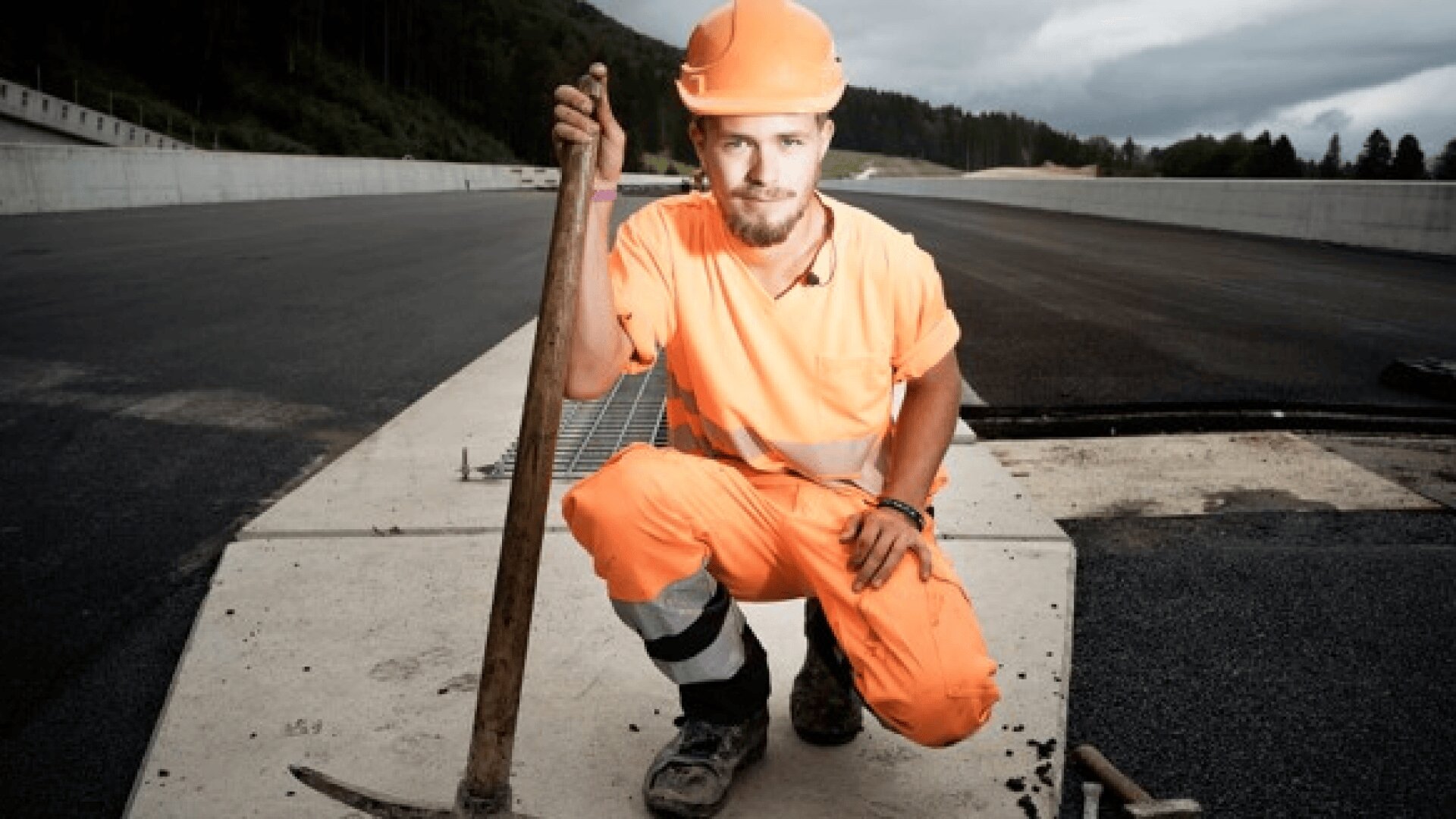 Ein Bauarbeiter in orangefarbener Sicherheitskleidung und mit Helm kniet mit einer Spitzhacke in der Hand auf einer frisch geteerten Straße, im Hintergrund Bäume und wolkenverhangener Himmel.