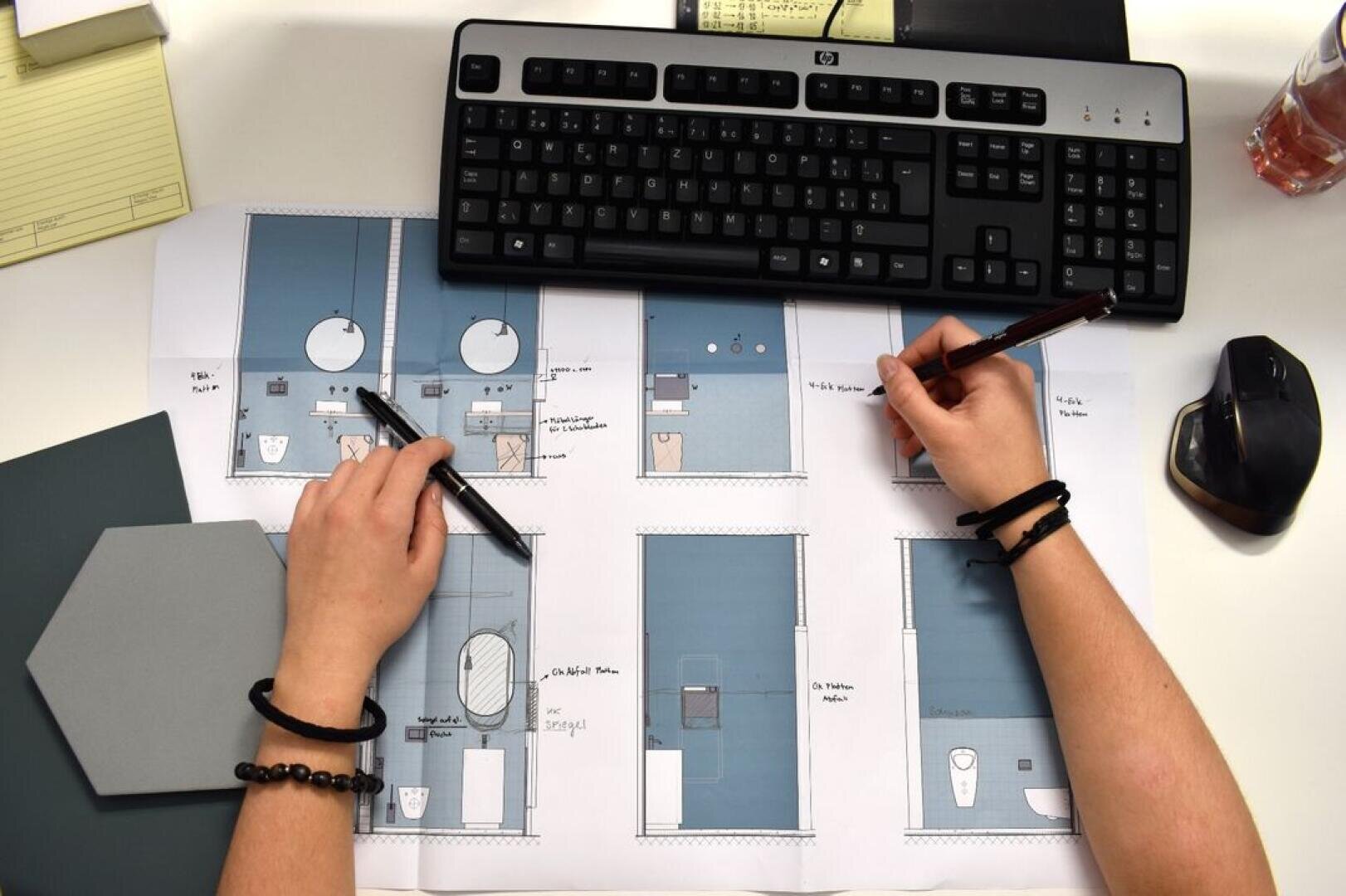 Eine Person macht Skizzen und Notizen zu architektonischen Badezimmergrundrissen an einem Schreibtisch mit einer Tastatur, einer Maus, einem Notizbuch und einem sechseckigen Kachelmuster.