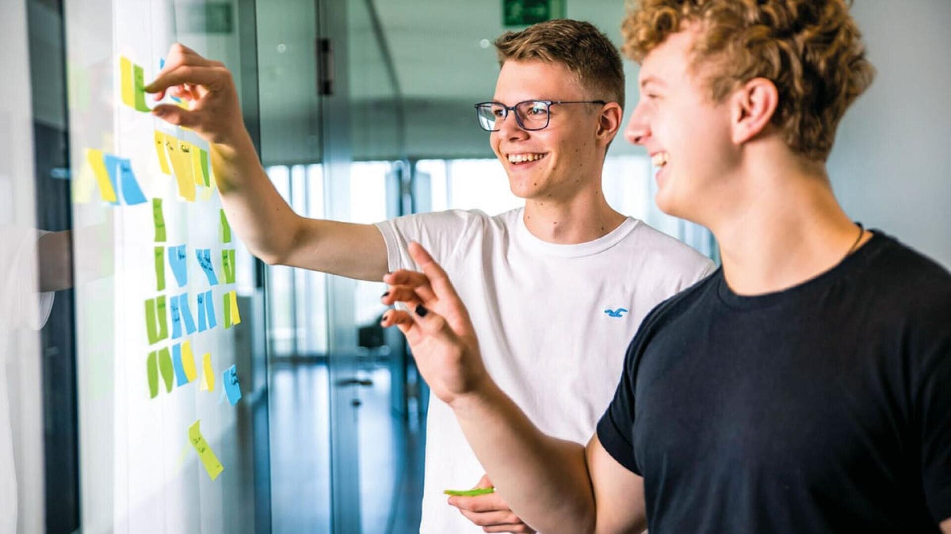 Zwei junge Männer lächeln und arbeiten zusammen, indem sie Klebezettel an einer Glaswand in einem hell erleuchteten modernen Bürokorridor anbringen.