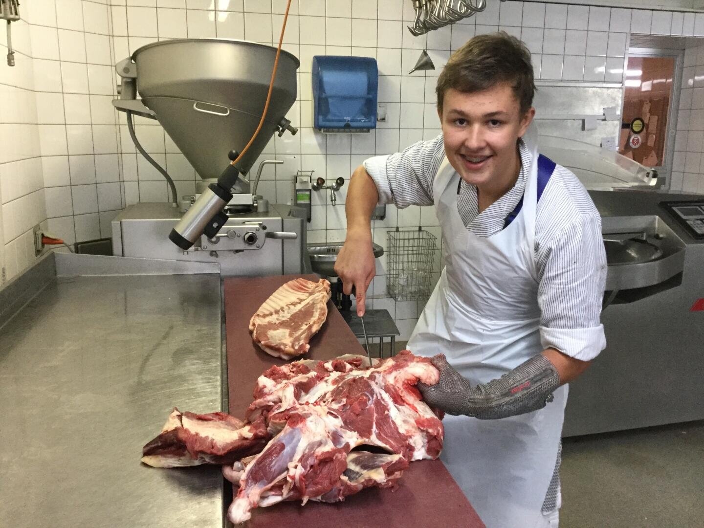 Ein lächelnder junger Metzger in gestreiftem Hemd und Schürze schneidet mit einem Messer rohes Fleisch auf einer Metalltheke in einem gekachelten Raum, wobei er an einer Hand einen Metallschutzhandschuh trägt.