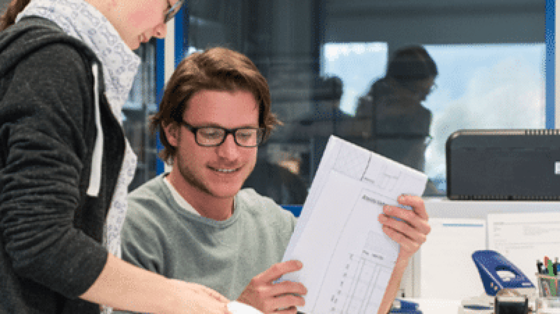 Zwei Personen in einem Büro sehen sich gemeinsam Dokumente an. Eine Person sitzt, lächelt und hält Papiere in der Hand, während die andere in der Nähe steht und auf die Dokumente zeigt. Im Hintergrund sind Büromaterial und ein Computer zu sehen.