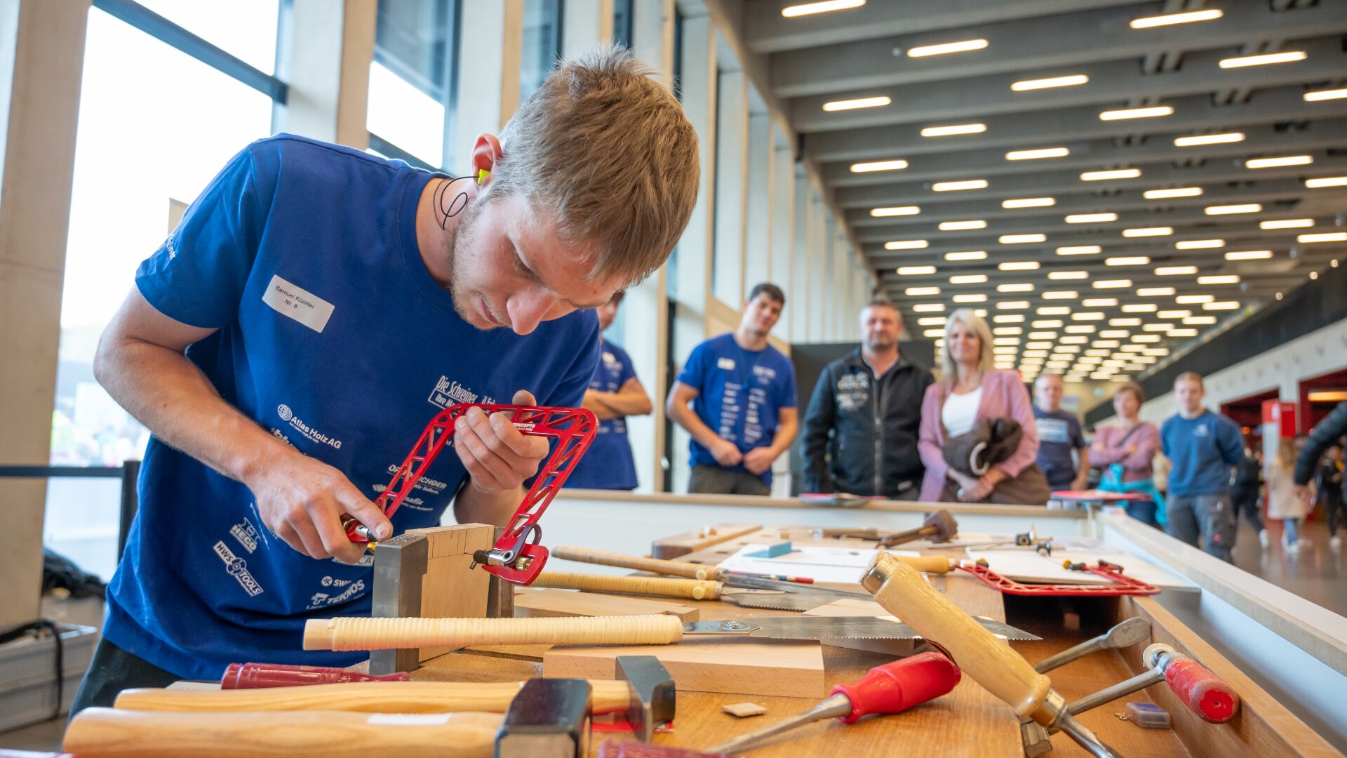 Ein junger Mann arbeitet mit einer Laubsäge an einem Tisch an einem Holzbearbeitungsprojekt, während in der Nähe mehrere Werkzeuge verstreut sind. Im Hintergrund sehen ihm Menschen in einem hell erleuchteten Innenraum zu.