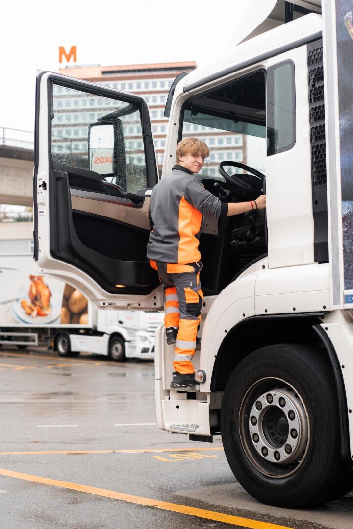 Eine Person in orangefarbener und grauer Arbeitskleidung klettert auf den Fahrersitz eines weißen Lastwagens, der auf einem Parkplatz geparkt ist, wobei im Hintergrund Gebäude und andere Lastwagen zu sehen sind.