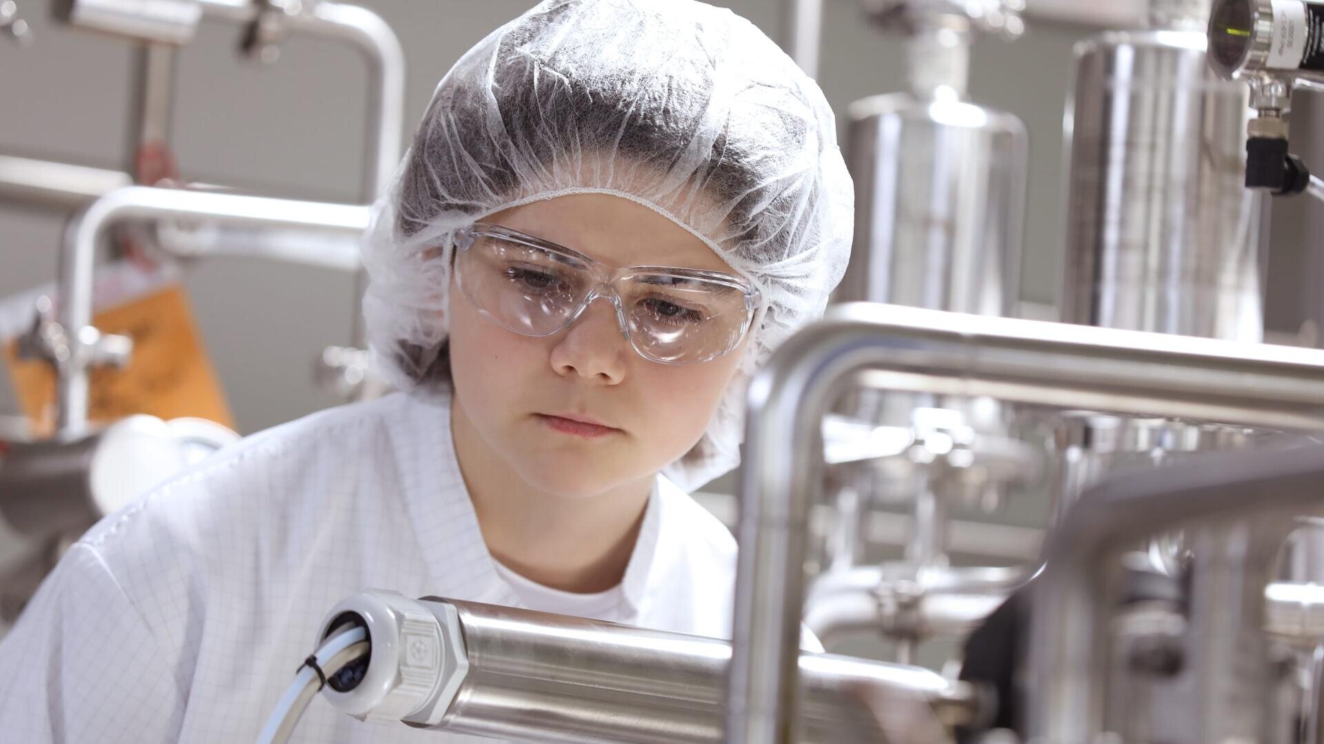 Eine Person, die ein Haarnetz, eine Schutzbrille und einen Laborkittel trägt, inspiziert in einem Labor oder einer Produktionsstätte Industrieanlagen aus rostfreiem Stahl.