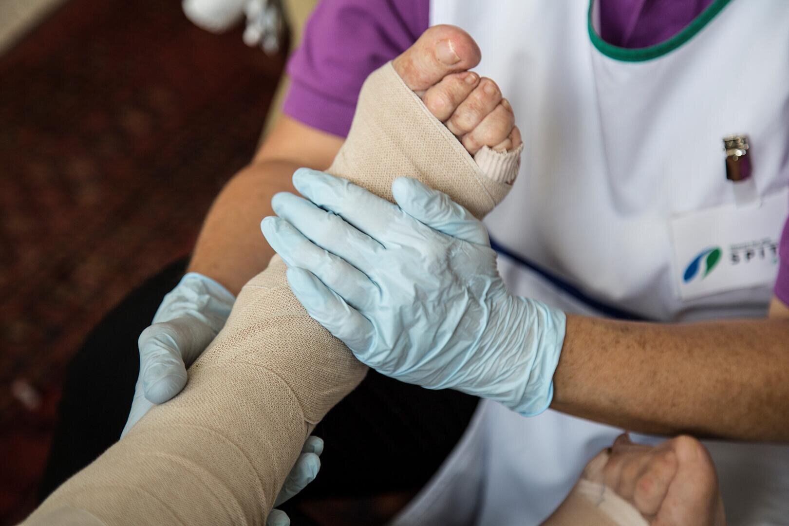 Eine Pflegekraft mit blauen Handschuhen wickelt einen Verband um Unterschenkel und Fuß einer Person und leistet medizinische Hilfe.