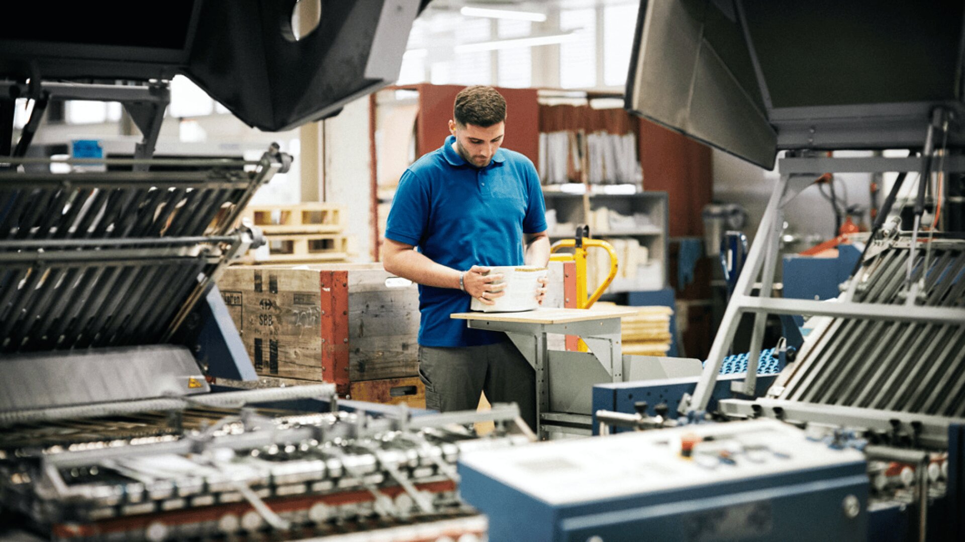 Ein Arbeiter in einem blauen Polohemd prüft einen Papierstapel in einer geschäftigen industriellen Druckerei, die von Druckmaschinen und Geräten umgeben ist.