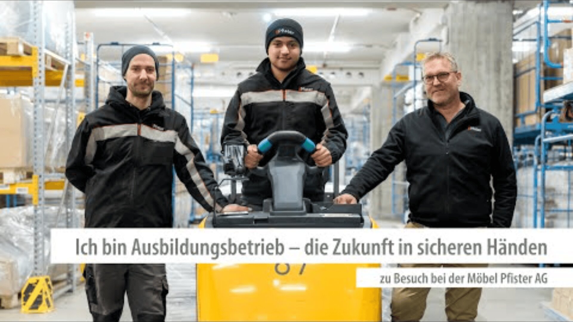 Drei Männer in Arbeitsuniformen stehen in einem Lagerhaus, wobei einer einen gelben Palettenheber bedient. Im Hintergrund sind Regale mit Kisten zu sehen. Der deutsche Text lautet: Ich bin Ausbildungsbetrieb - die Zukunft in sicheren Händen.
