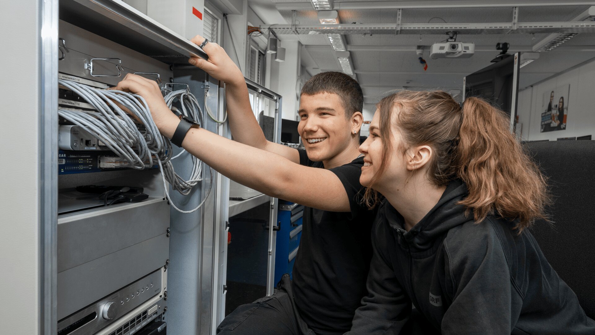 Zwei junge Menschen, ein Mann und eine Frau, lächeln, während sie gemeinsam an einem Serverschrank arbeiten und Netzwerkkabel in einer technischen oder Computerlaborumgebung anschließen.