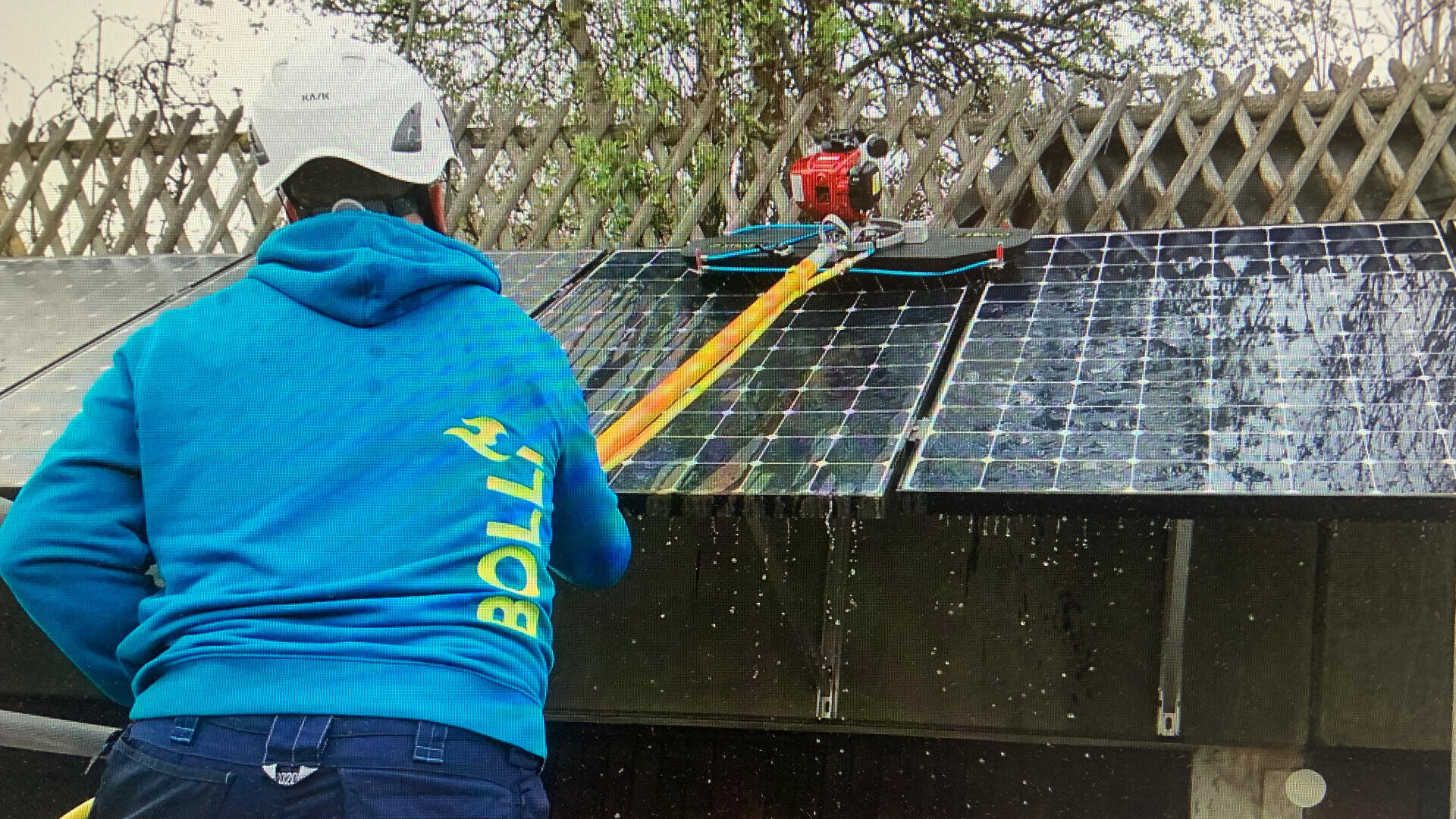 Eine Person mit blauem Kapuzenpulli und weißem Helm verwendet ein langes Werkzeug, um eine Reihe von Solarzellen auf dem Dach zu reinigen, wobei Wasser an den Platten herunterläuft. Im Hintergrund sind Holzzäune und Bäume zu sehen.