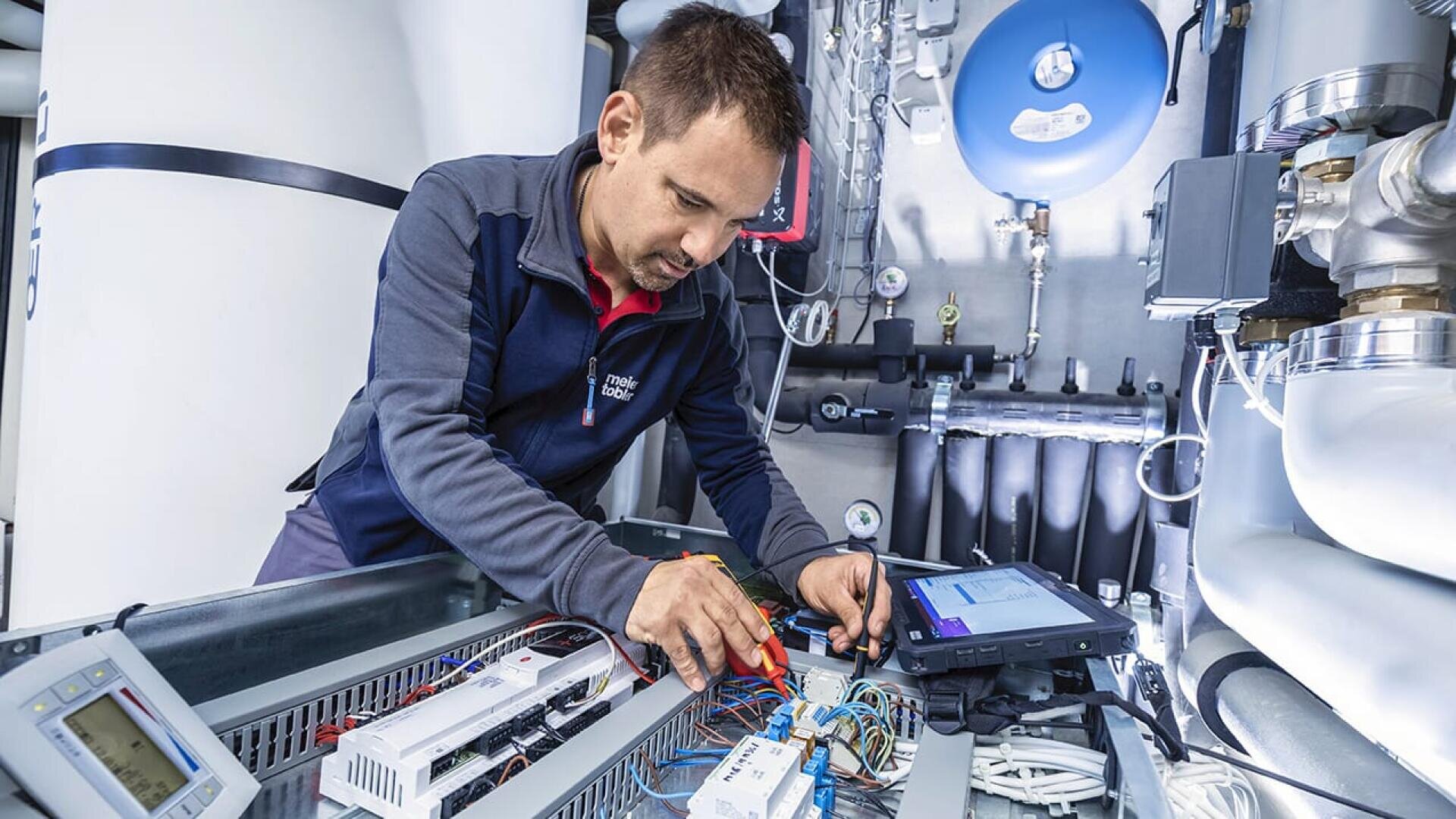 Ein Techniker in einer blauen Jacke inspiziert und repariert mit Werkzeugen elektrische Komponenten in einer industriellen Schalttafel, umgeben von Drähten, Rohren und Überwachungsgeräten in einem hell erleuchteten Technikraum.