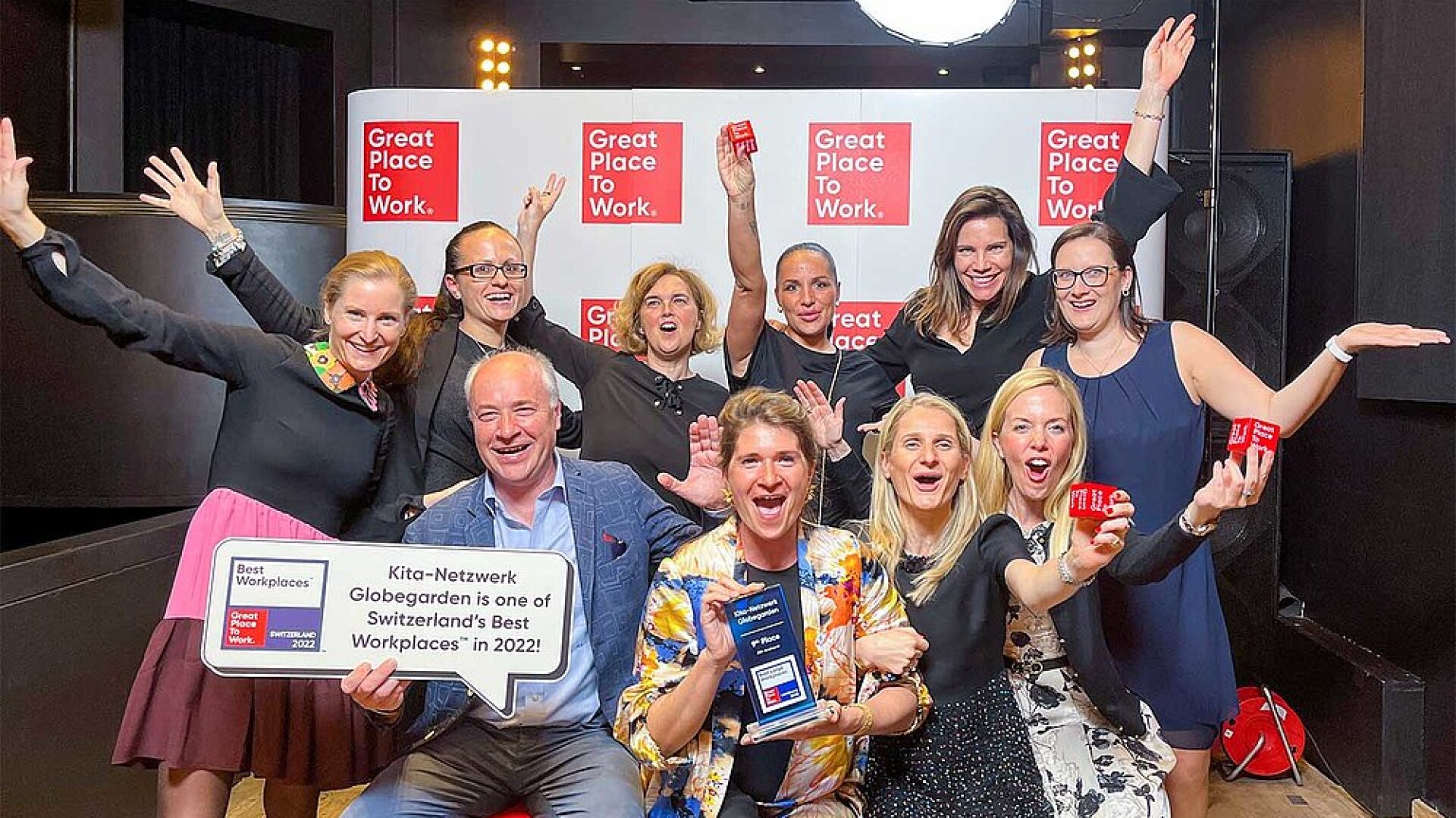 Eine fröhliche Gruppe von Menschen posiert mit Auszeichnungen und Zertifikaten in der Hand vor einer Wand mit mehreren Great Place to Work-Schildern. Eine Person hält ein Plakat, das das Unternehmen als Top-Arbeitsplatz in der Schweiz auszeichnet.
