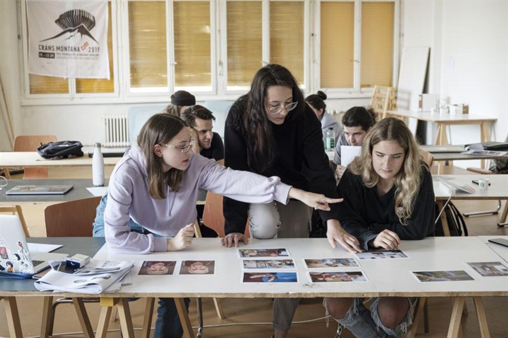 Drei junge Frauen sitzen und stehen um einen mit Fotos bedeckten Tisch herum, diskutieren und zeigen auf die Bilder. Andere Schüler arbeiten im Hintergrund in einem Klassenzimmer mit großen Fenstern und einem Crans-Montana-Banner an der Wand.