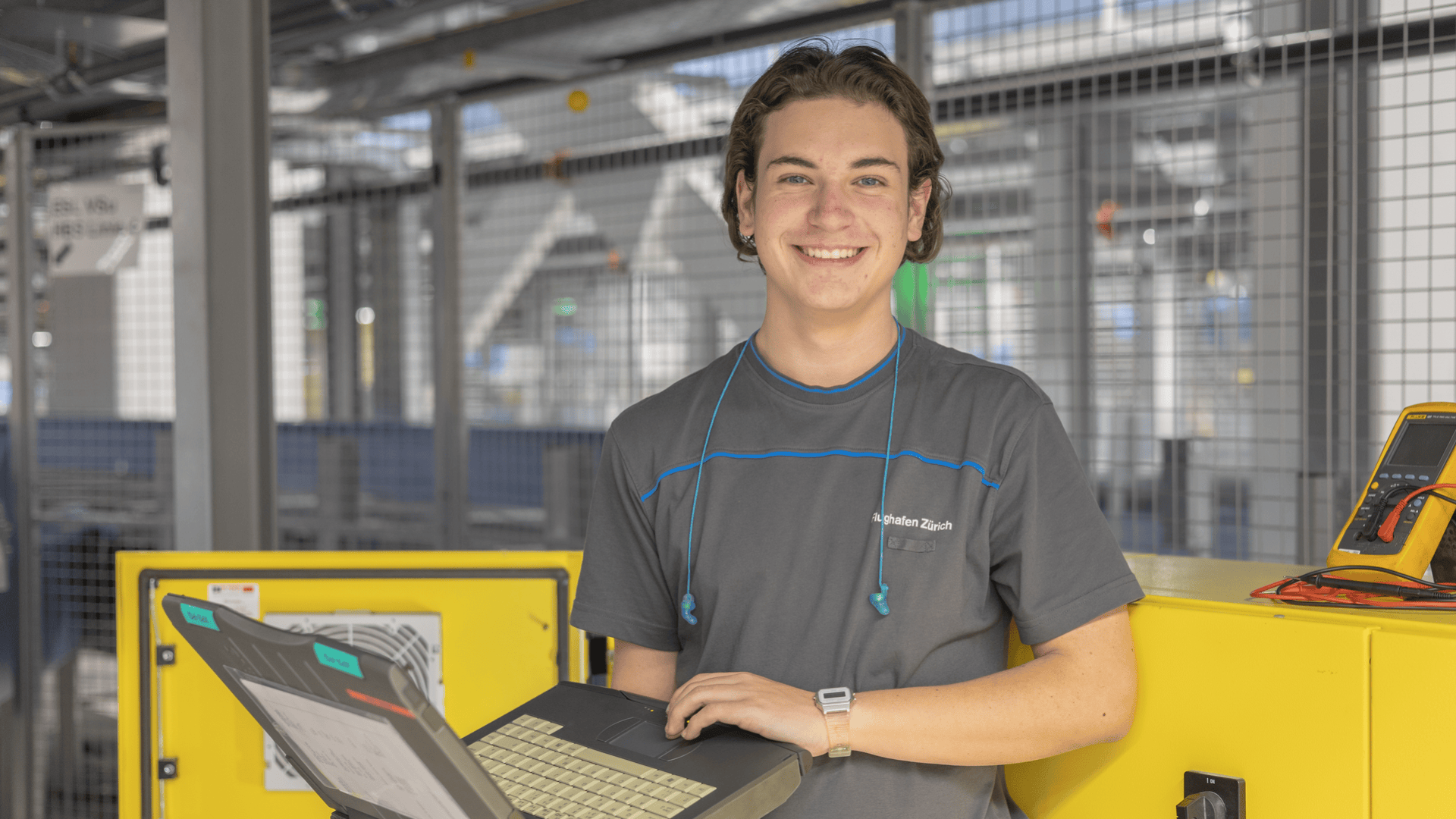 Eine junge Person in einem grauen Hemd steht im Haus, lächelt und hält einen aufgeklappten Laptop in der Hand. Sie hat einen blaugrünen Kopfhörer um den Hals, und im Hintergrund sind gelbe Gerätekästen und ein digitales Messgerät zu sehen.