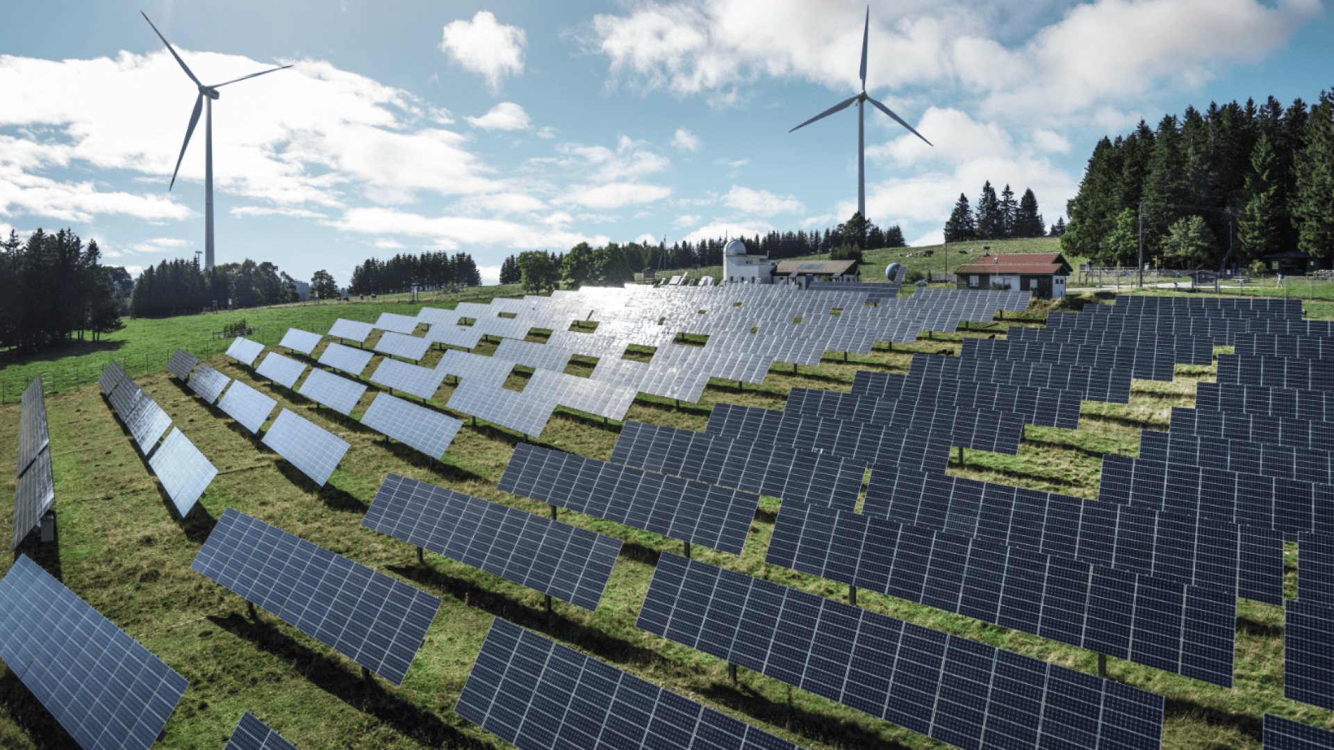 Ein großer Solarpark auf einer grünen Wiese mit Windturbinen im Hintergrund unter einem teilweise bewölkten Himmel, umgeben von Bäumen und einigen kleinen Gebäuden.
