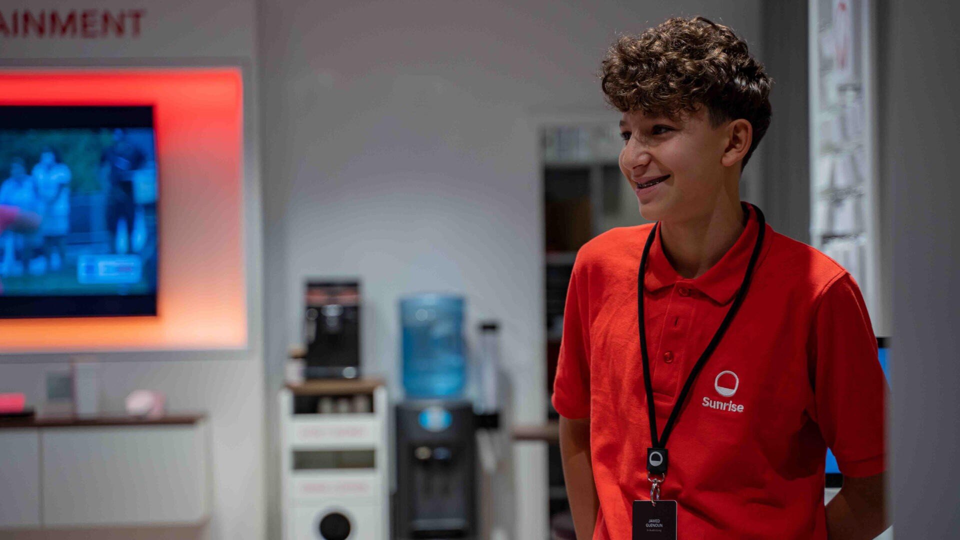 Eine junge Person mit lockigem Haar, die ein rotes Sunrise Polohemd und ein Schlüsselband trägt, lächelt, während sie in einem modernen Geschäft mit einem Fernseher und einem Wasserspender im Hintergrund steht.