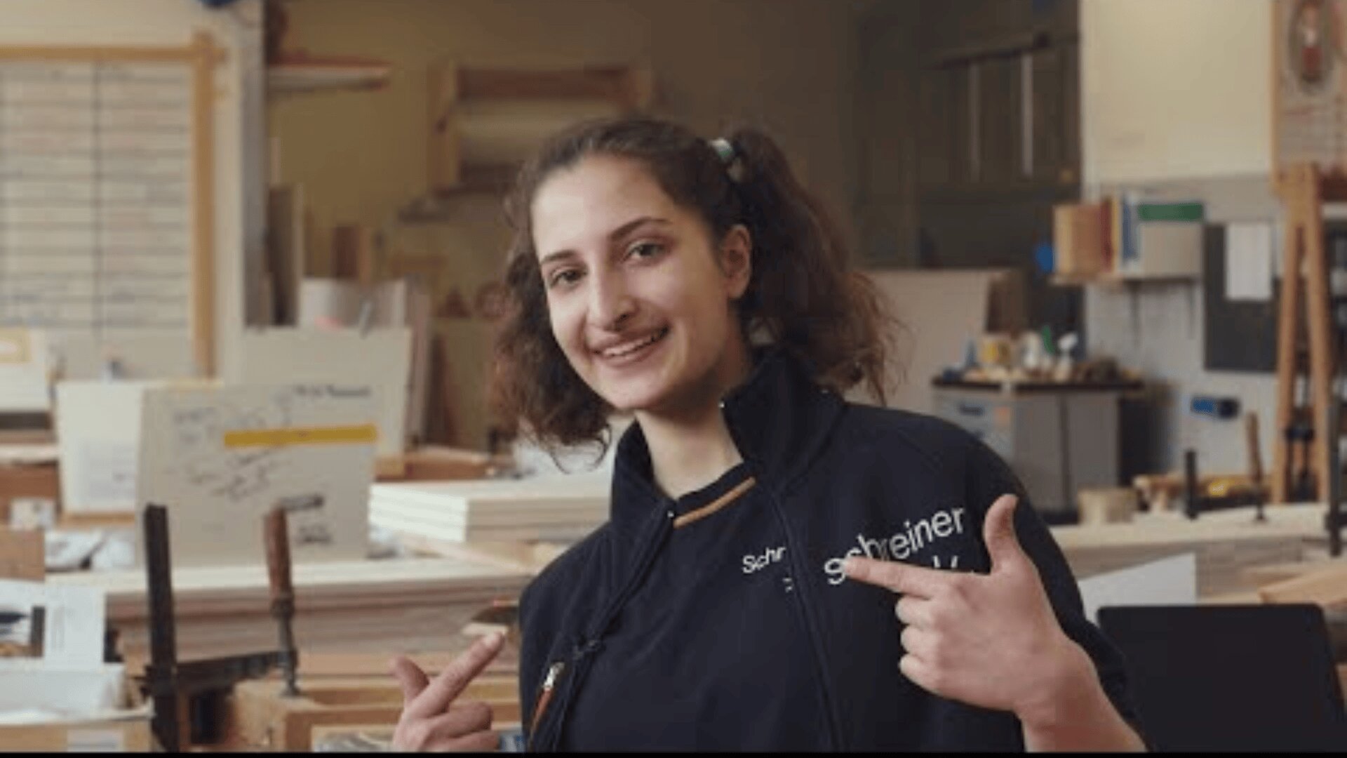 Eine junge Frau mit braunen Haaren in Zöpfen lächelt und zeigt auf ihre Arbeitsjacke in einer Tischlerei oder Schreinerei, umgeben von Werkzeugen, Holz und Werkbänken.
