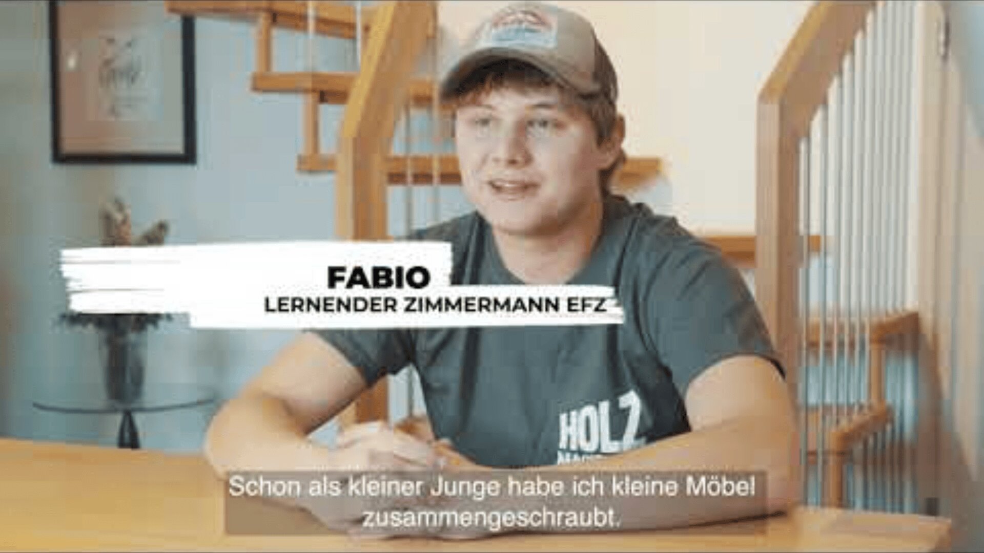 Ein junger Mann mit Mütze und HOLZ-Hemd sitzt an einem Holztisch in der Nähe einer Treppe. Der Text auf dem Bildschirm lautet: FABIO, LERNENDER ZIMMERMANN EFZ. Untertitel lauten: Schon als kleiner Junge habe ich kleine Möbel zusammengeschraubt.