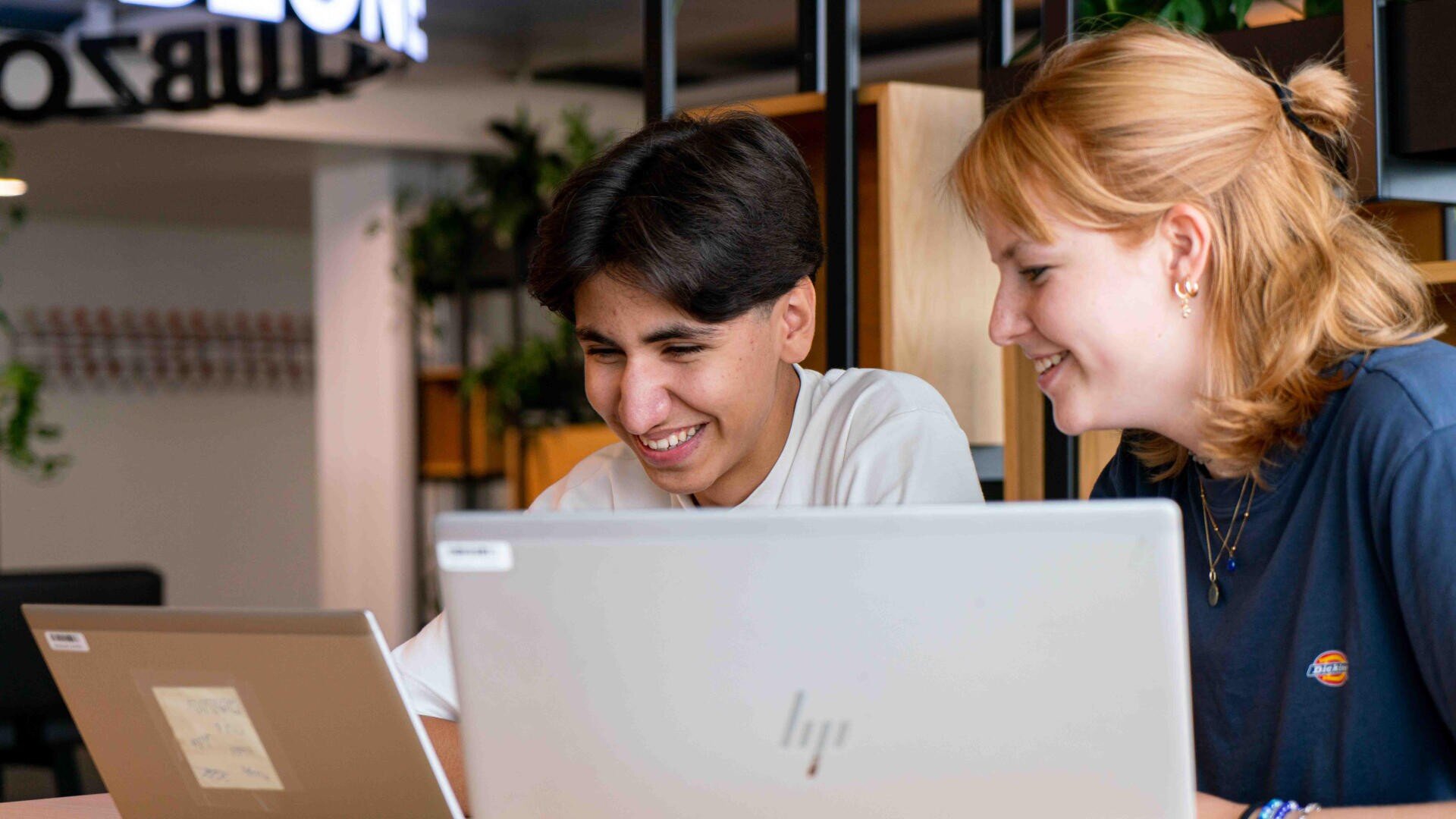Zwei junge Leute sitzen nebeneinander an einem Tisch, lächeln und schauen auf ihre Laptops in einem modernen, hell erleuchteten Arbeitsbereich mit Pflanzen und Regalen im Hintergrund.