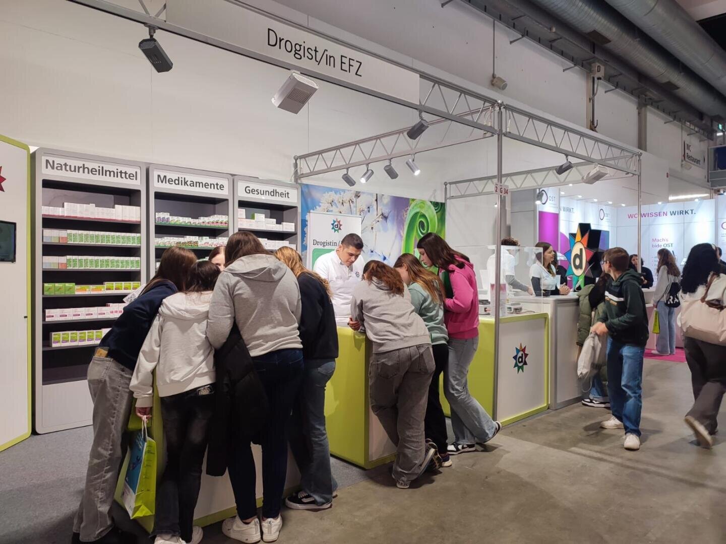 Eine Gruppe von Personen steht an einem hellen Messestand mit der Aufschrift "Drogist/in EFZ", mit Regalen voller Produkte und Informationsdisplays, während das Personal hinter dem Tresen die Besucher in einer belebten Ausstellungshalle berät.