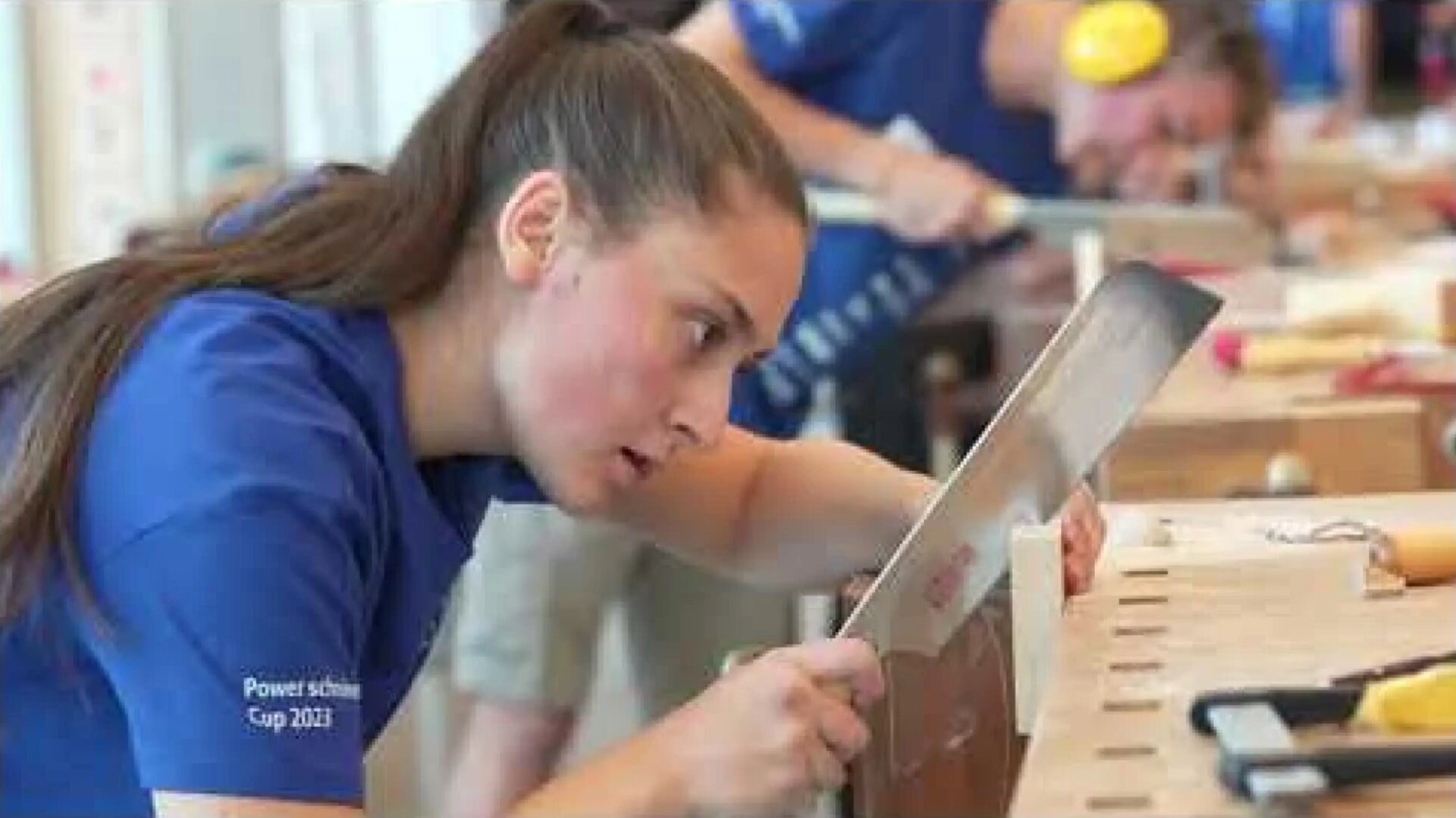 Eine konzentrierte Frau in einem blauen Hemd sägt mit einer Handsäge Holz, während eine andere Person im Hintergrund arbeitet. Beide nehmen an einem Holzbearbeitungswettbewerb teil.