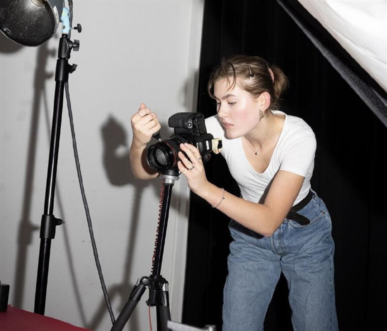 Eine junge Frau in weißem T-Shirt und Jeans stellt in einem Fotostudio eine Kamera auf einem Stativ ein und bereitet sich konzentriert auf eine Aufnahme vor.