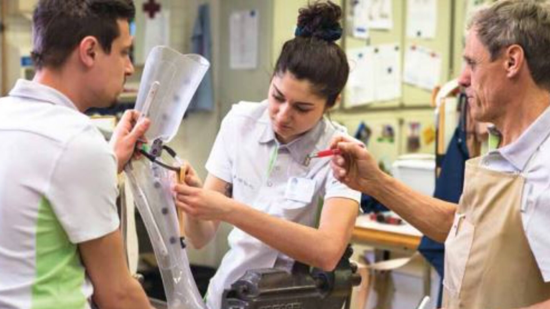 Drei Personen arbeiten zusammen in einer Werkstatt und passen eine durchsichtige Beinprothese an. Zwei von ihnen tragen weiße Uniformen, einer eine beige Schürze. Sie benutzen Werkzeuge und scheinen sich auf ihre Aufgabe zu konzentrieren, umgeben von technischen Zeichnungen und Werkzeugen.