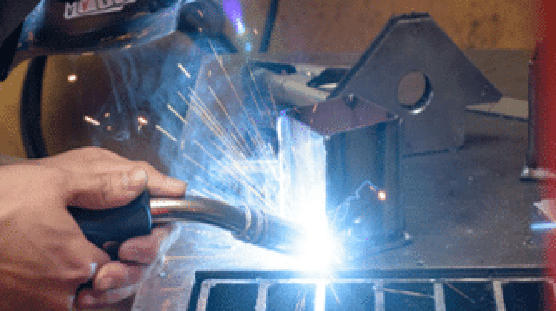Eine Person, die Schutzkleidung trägt, schweißt ein Metallteil auf einer Werkbank, wobei an der Kontaktstelle helle Funken und ein blaues Glühen entstehen.