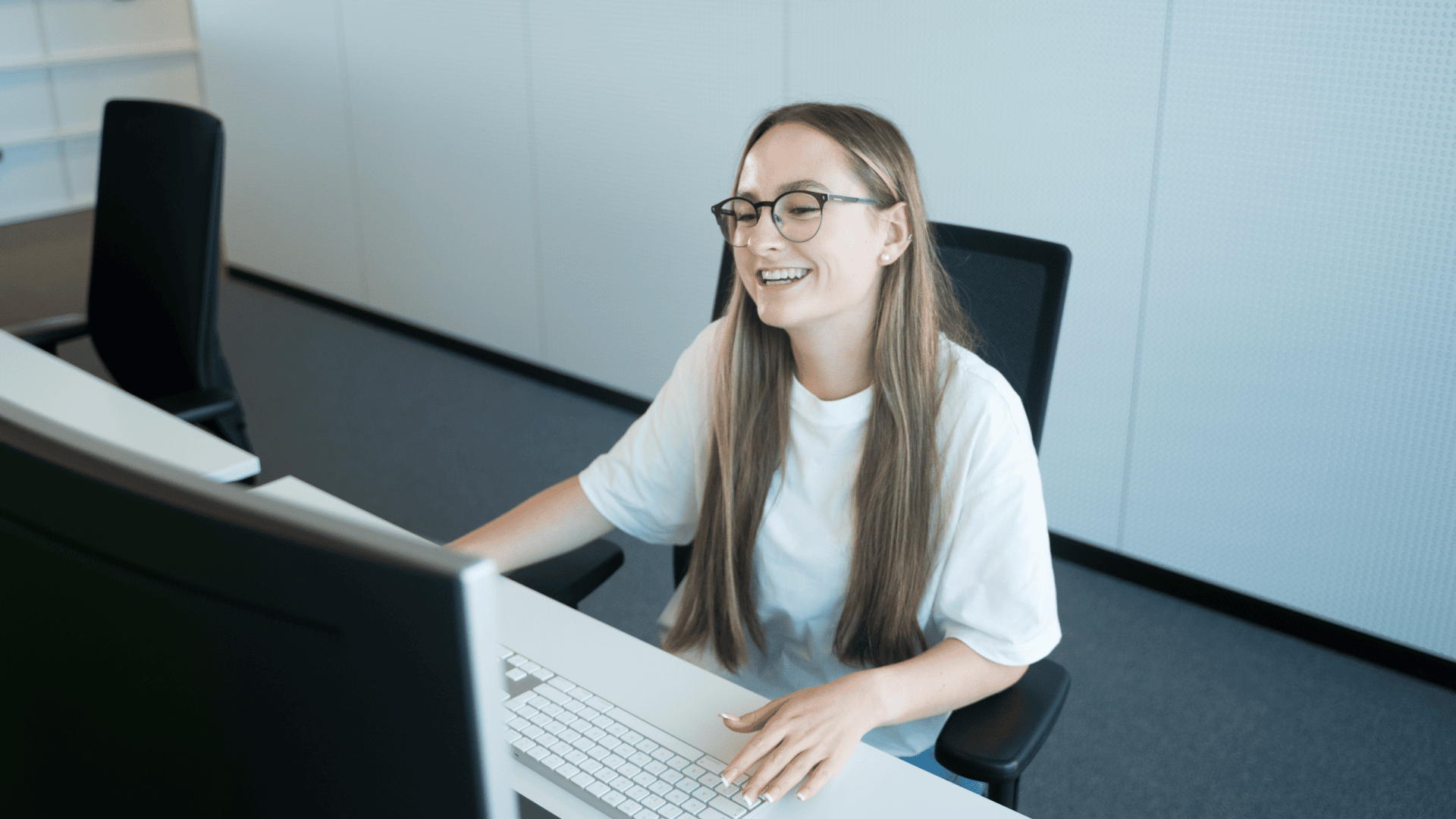 Eine junge Frau mit langen braunen Haaren und Brille lächelt, während sie an einem Schreibtisch sitzt und einen Computer in einer modernen Büroumgebung benutzt. Sie scheint entspannt und mit ihrer Arbeit beschäftigt zu sein.