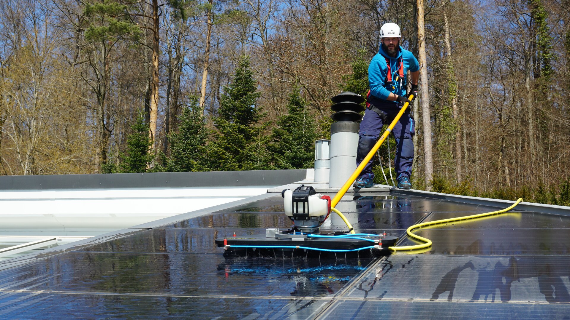 Ein Arbeiter in Schutzkleidung reinigt mit einem langstieligen Werkzeug Solarpaneele auf einem Dach, umgeben von Bäumen und blauem Himmel im Hintergrund.
