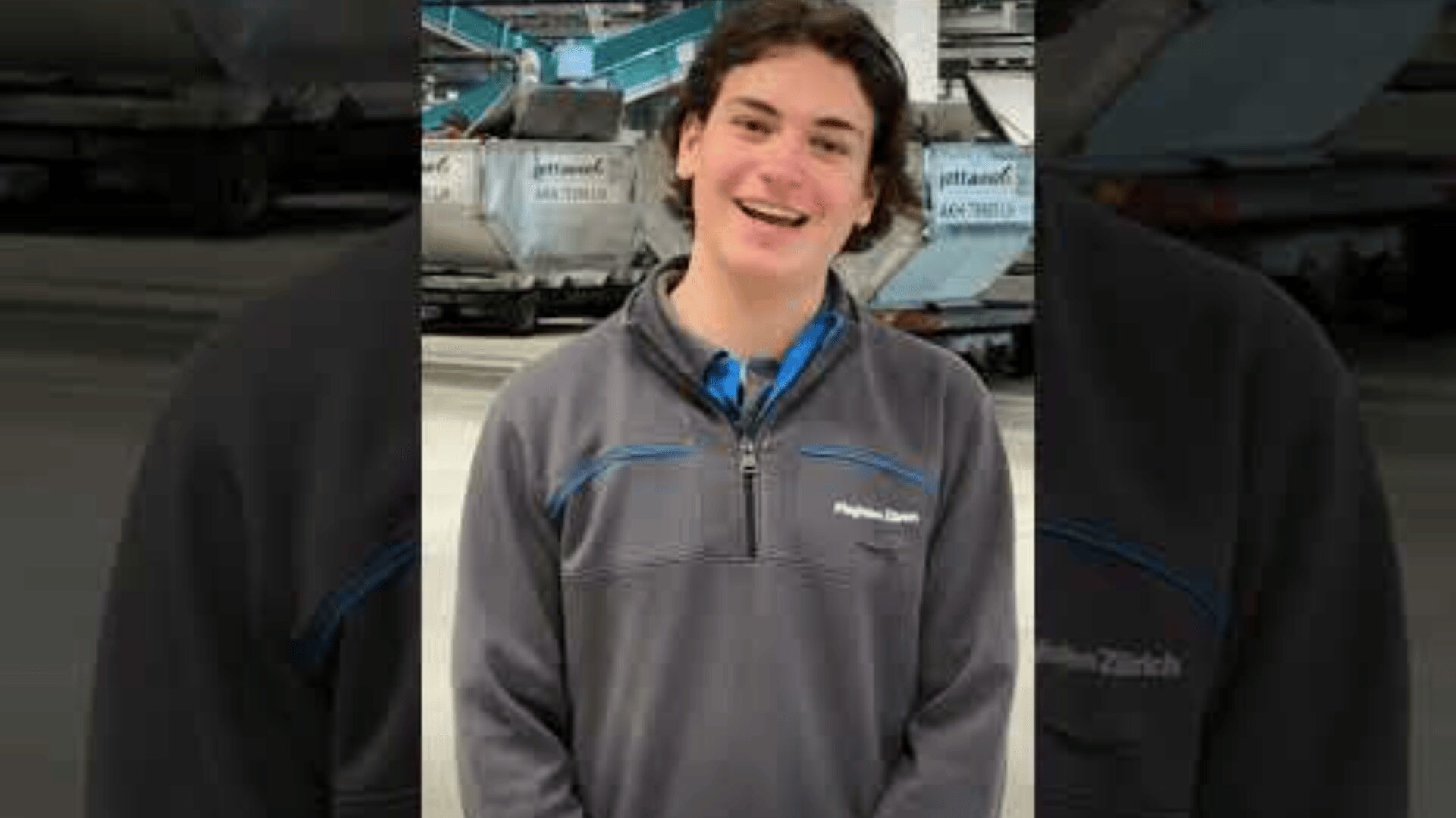 Eine Person mit langen braunen Haaren, die eine graue Jacke mit Reißverschluss trägt, lächelt, während sie in einem Haus steht. Im Hintergrund sind große Industrieanlagen und Maschinen zu sehen.